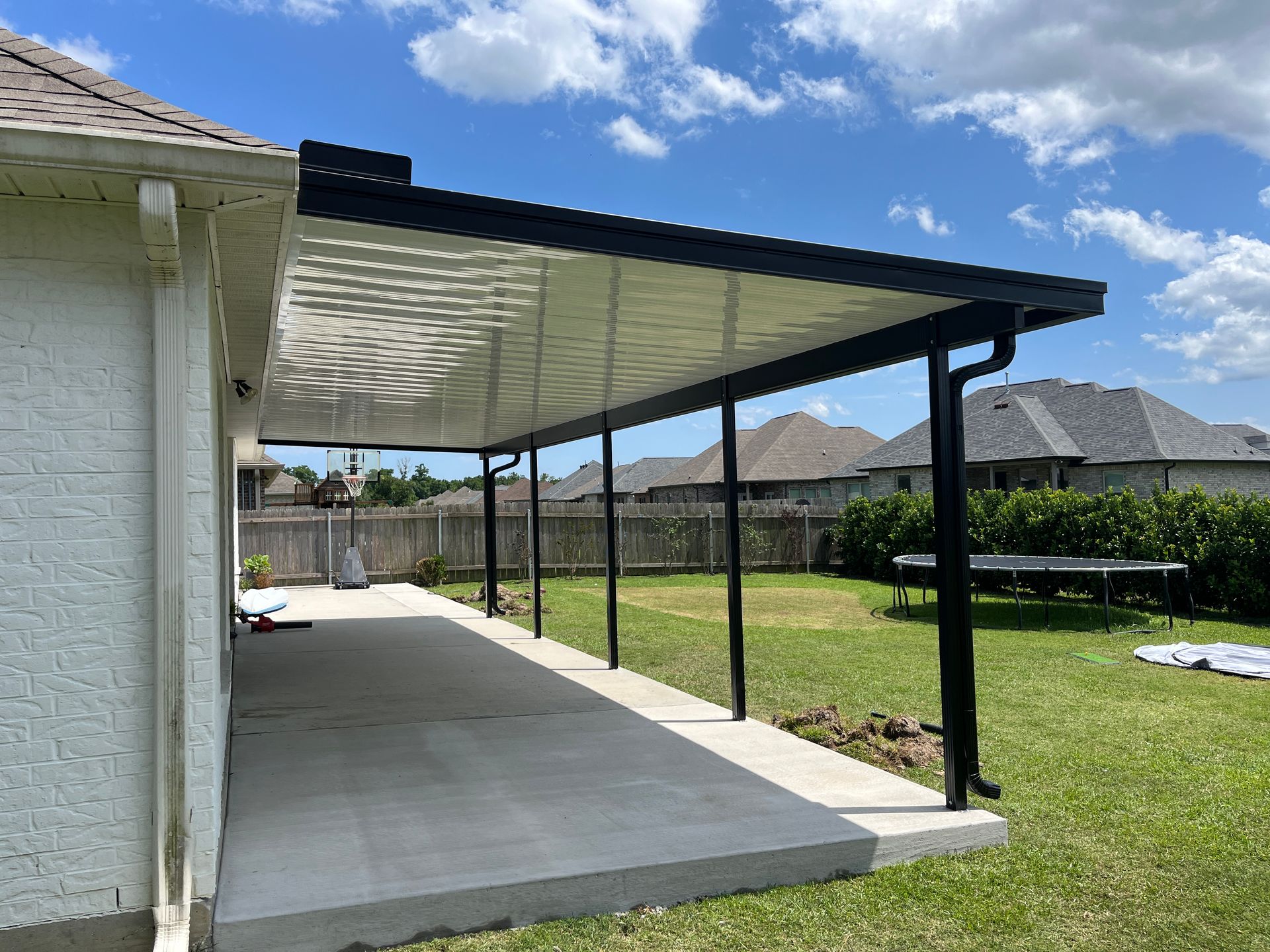 A backyard patio with a white textured roof cover supported by black posts, attached to a light-colored brick house.