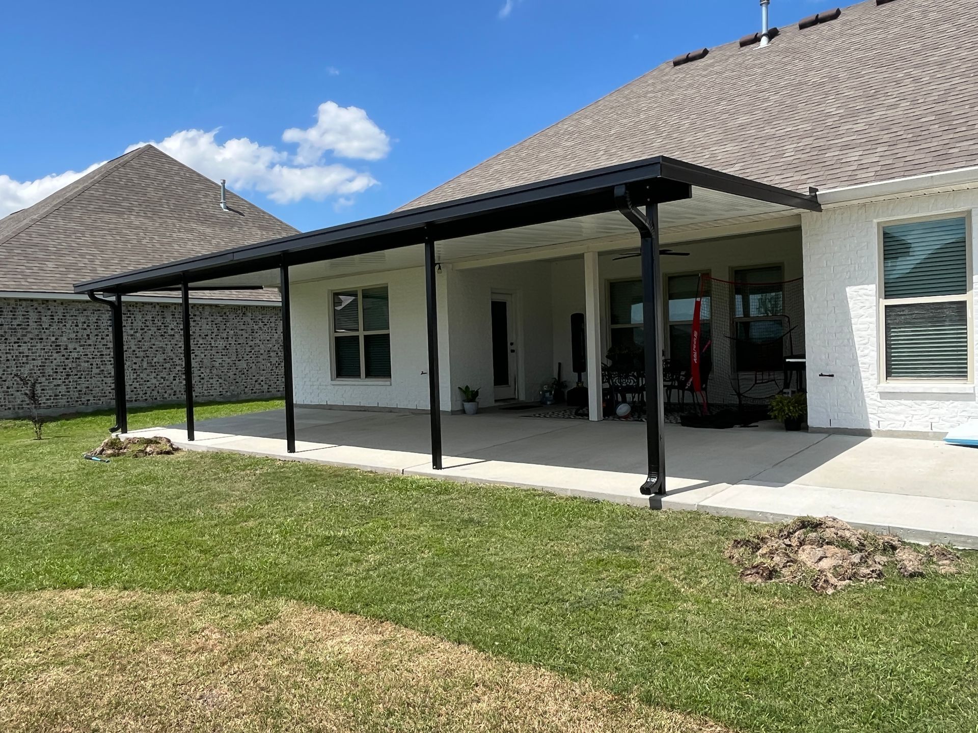 A white house features a long, black-framed patio cover extending over a concrete slab above a backyard lawn.