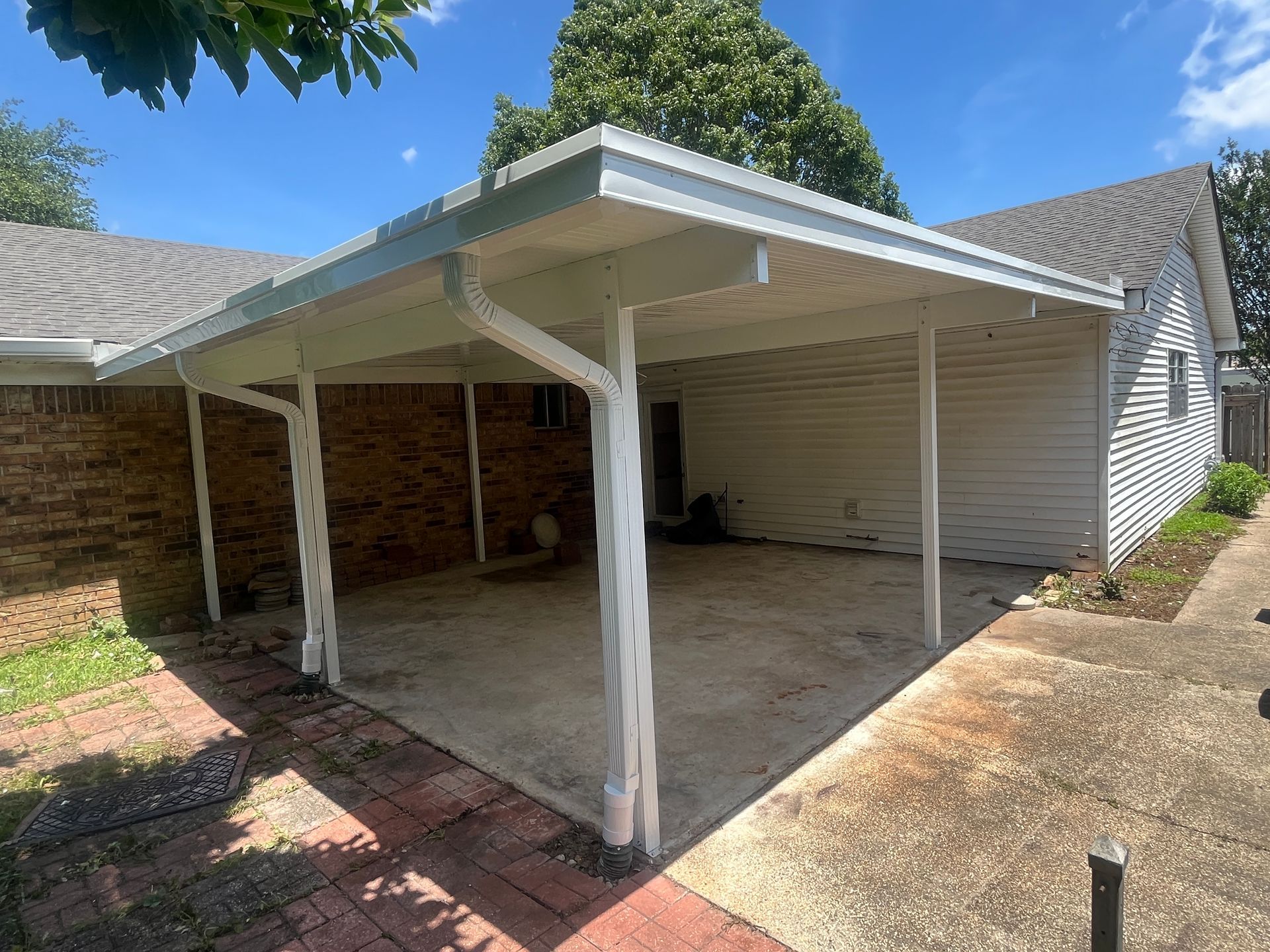 A covered patio or carport area with a concrete floor, white support pillars, and white siding attached to a brick house.