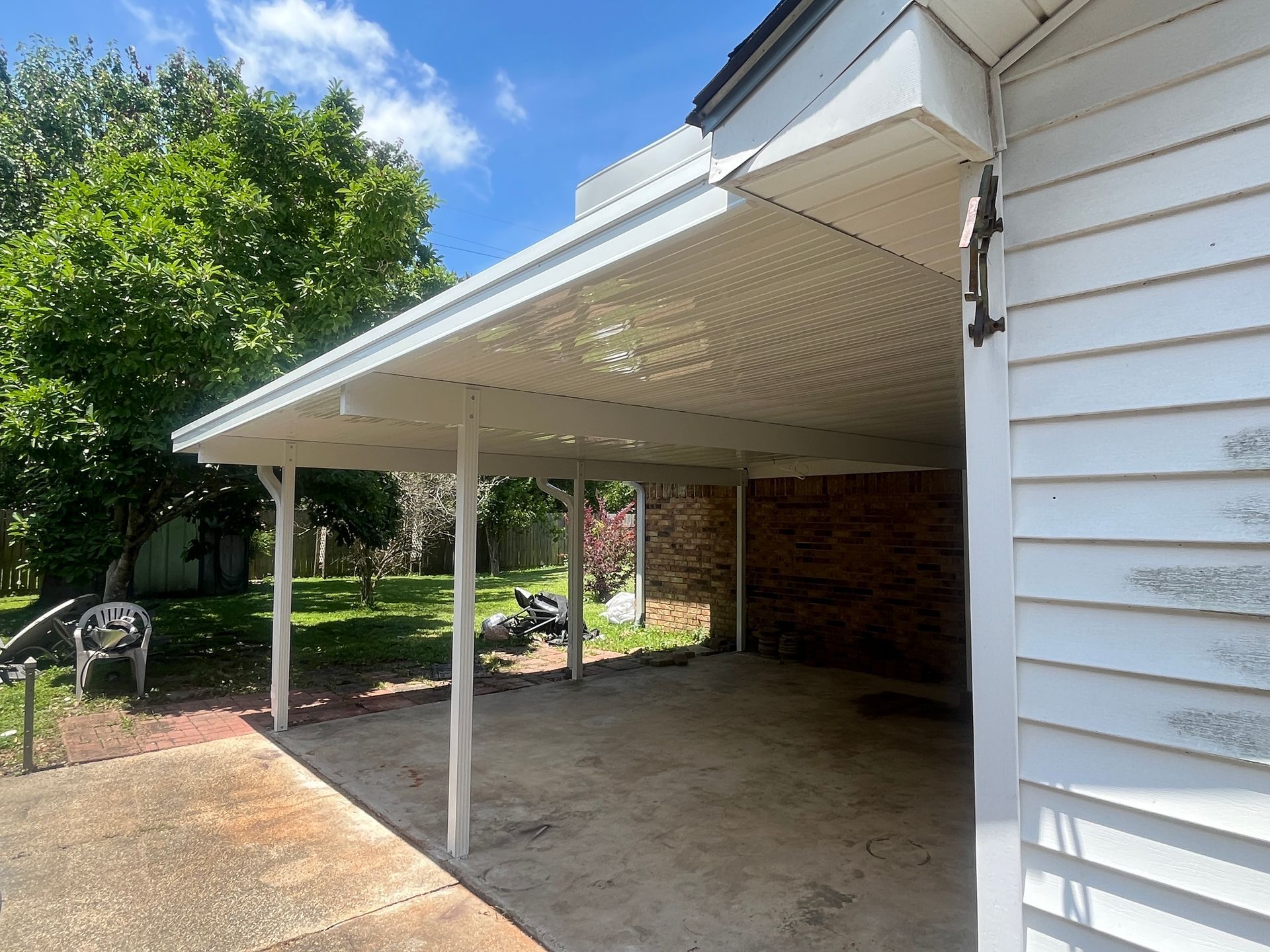 A white, wooden-sided house with a large, covered carport attached to its side, opening onto a concrete driveway.