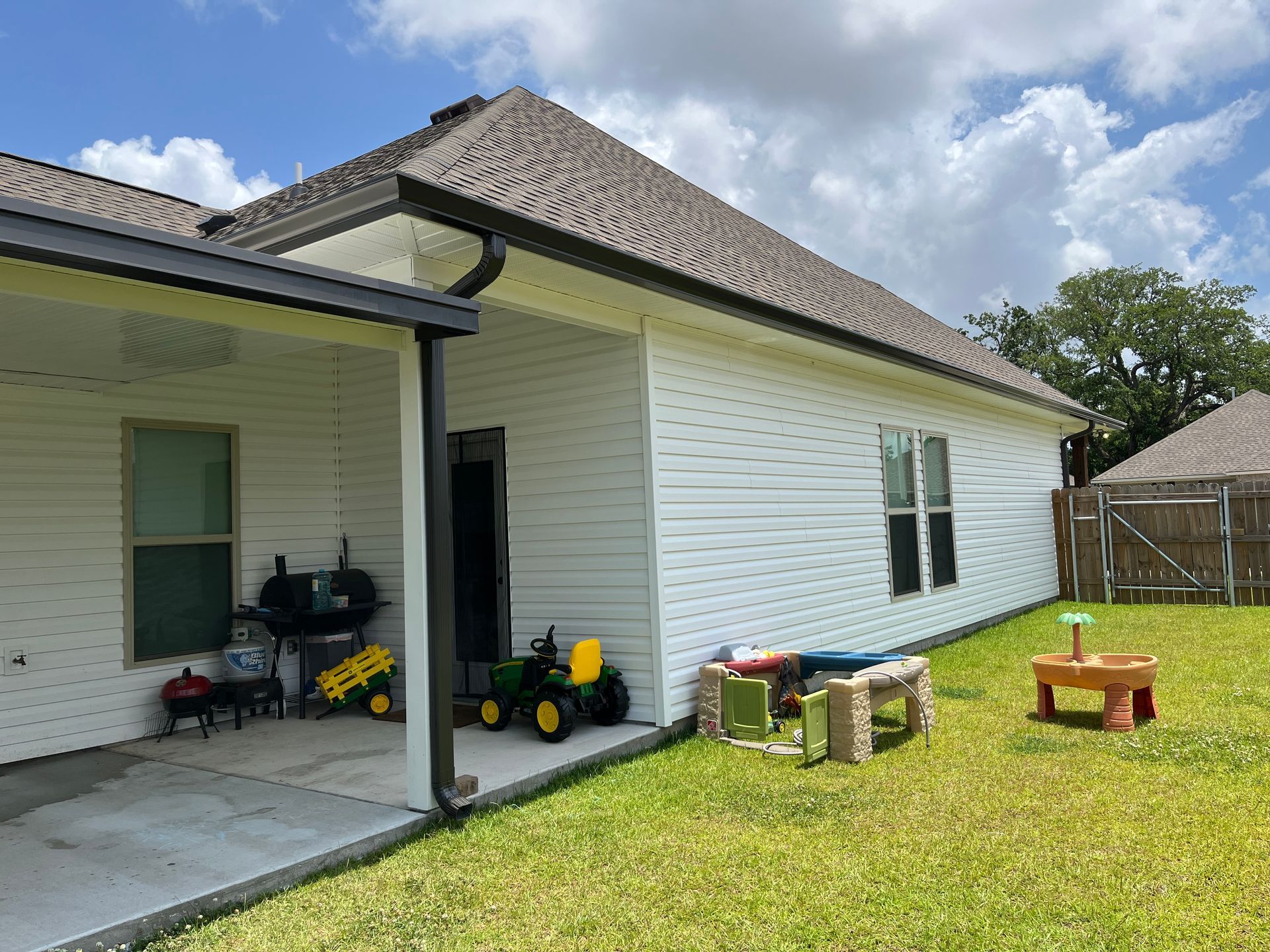 A covered backyard patio with a grill, a toy tractor, and play equipment, adjacent to a white house with a lawn.