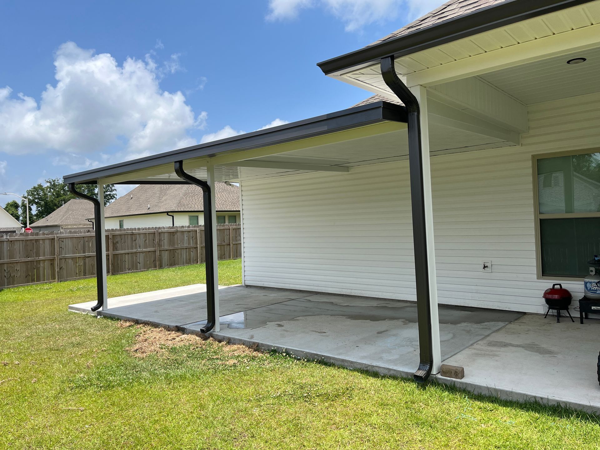 A side view of a house exterior featuring a white-sided wall, a concrete patio, and a covered roof with black gutters.