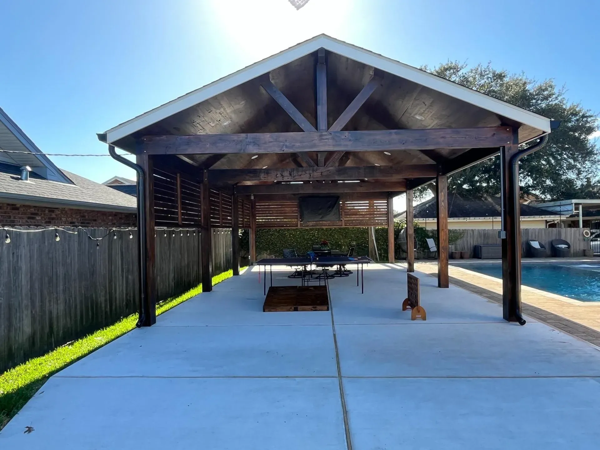 Covered backyard patio with wooden posts, exposed trusses, and a concrete floor beside a fence and pool.