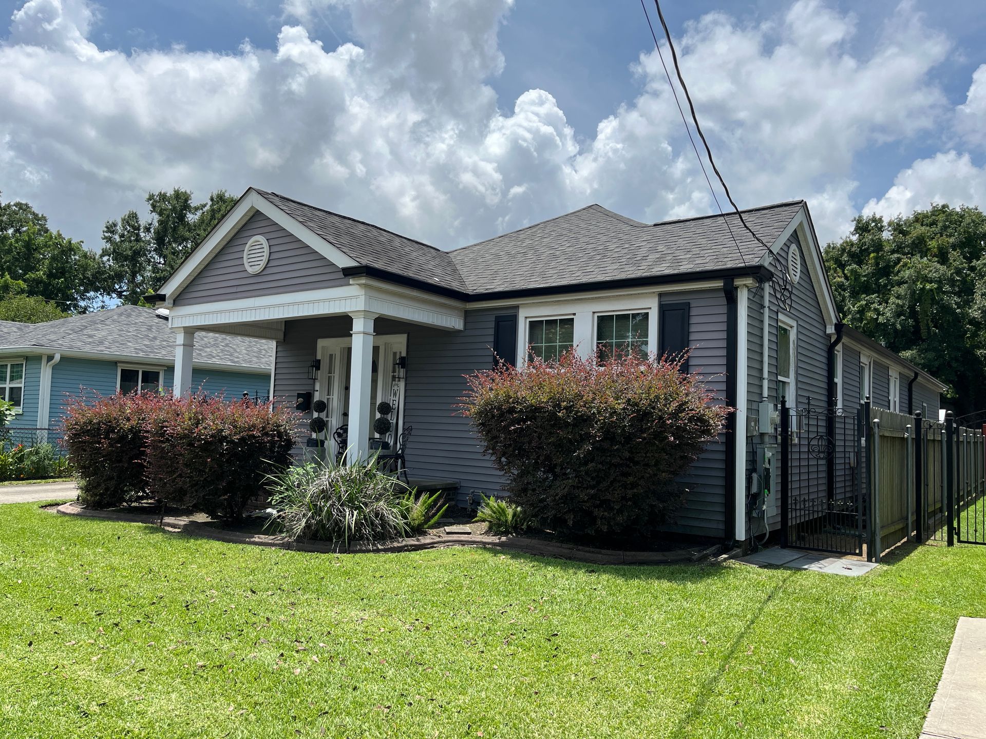 A gray, single-story house with a front porch, white columns, and a landscaped yard under a cloudy blue sky.