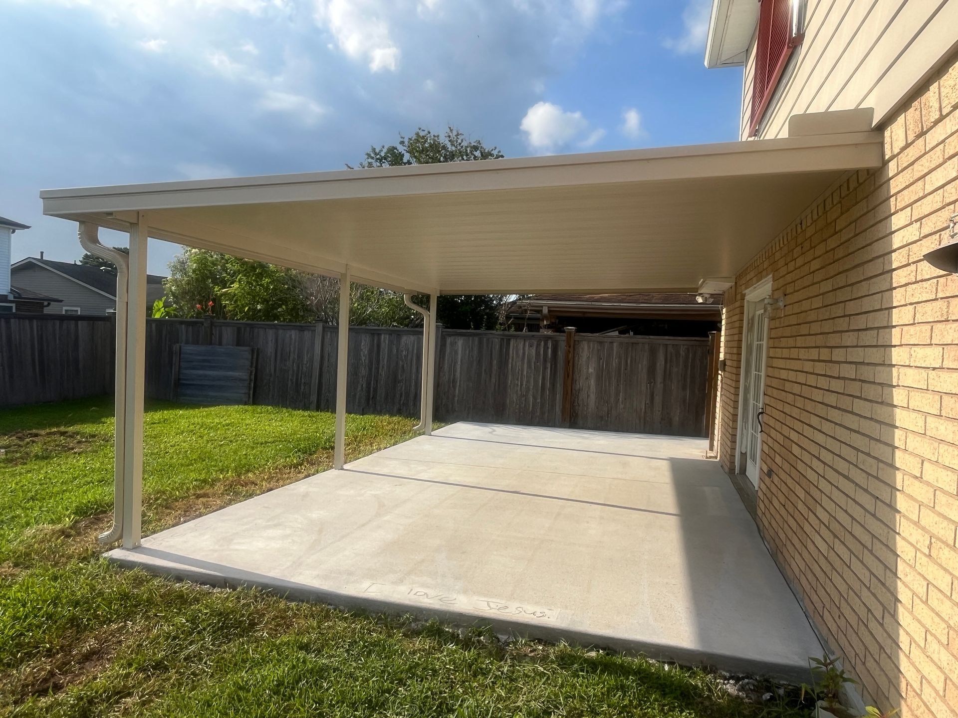 A cream-colored patio cover extends from a brick house wall over a concrete patio in a backyard with a wooden fence.