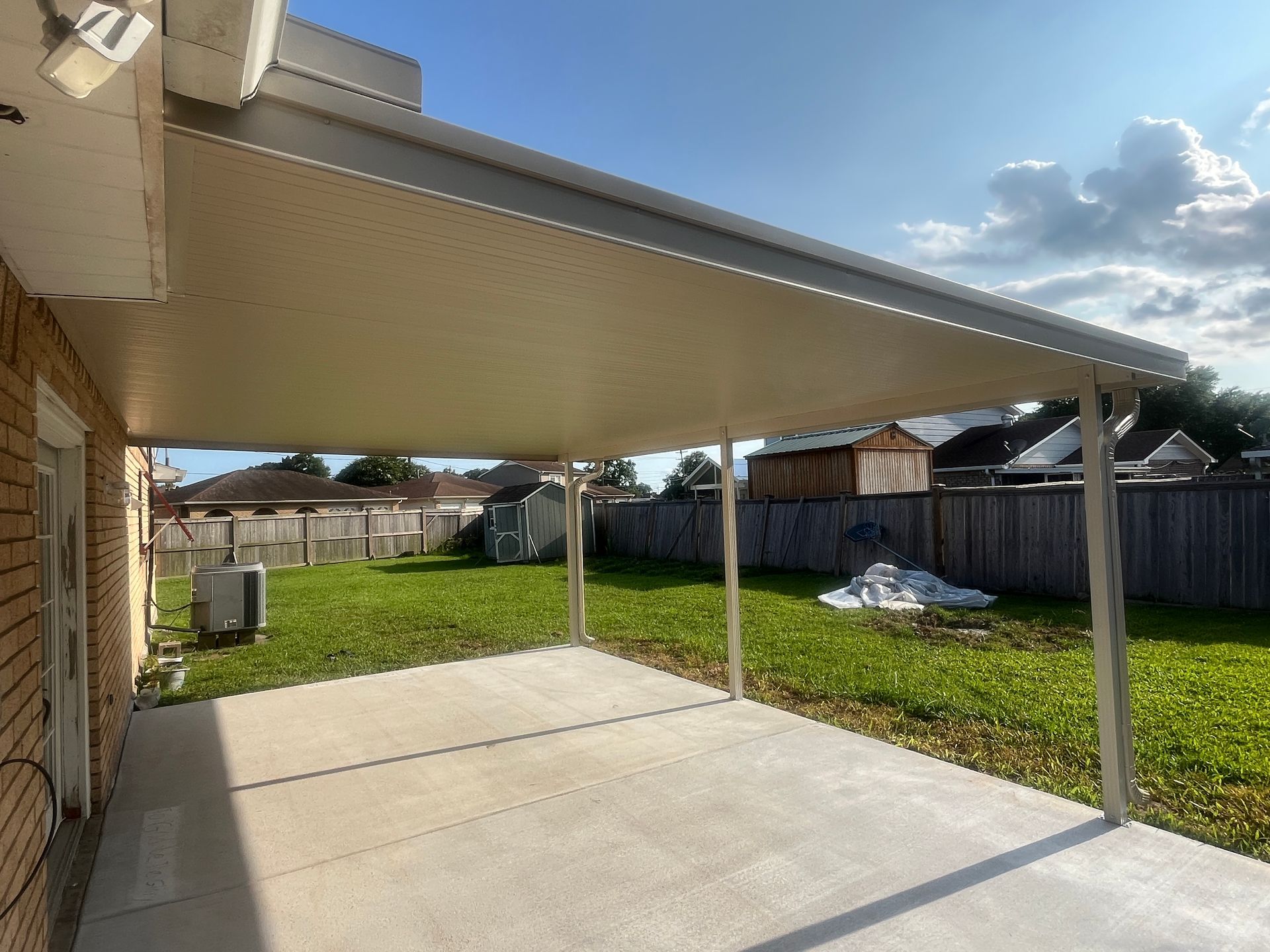 A concrete patio covered by an aluminum awning, looking out onto a grassy backyard with a wooden fence.
