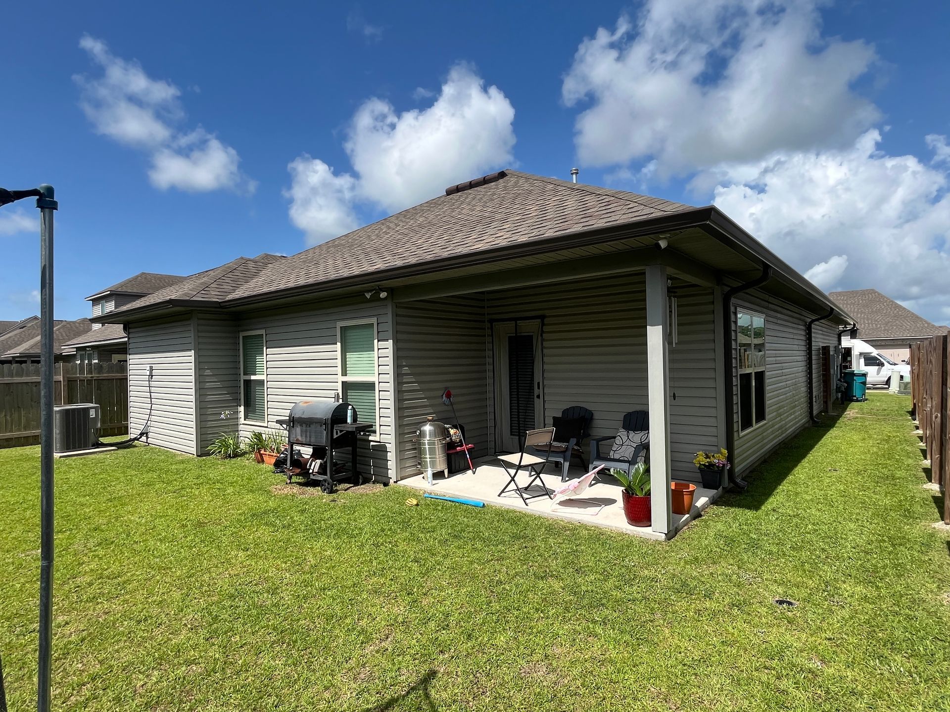 A backyard view of a light-colored, single-story house with a covered patio, a grill, and lawn furniture.