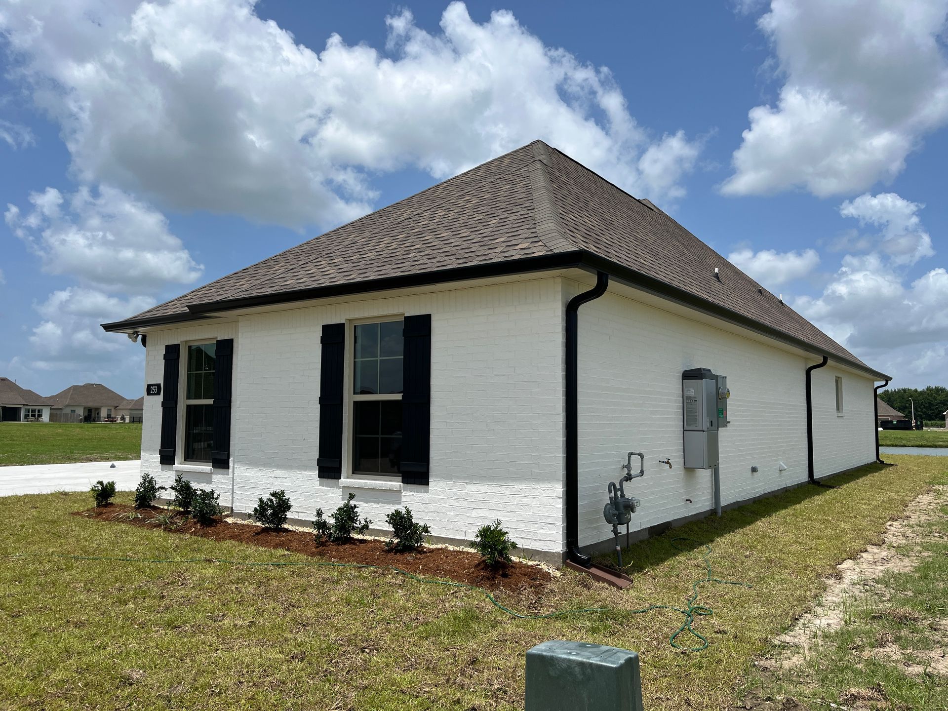 A white, single-story brick house with black shutters and trim sits on a grassy lot under a bright, cloudy sky.