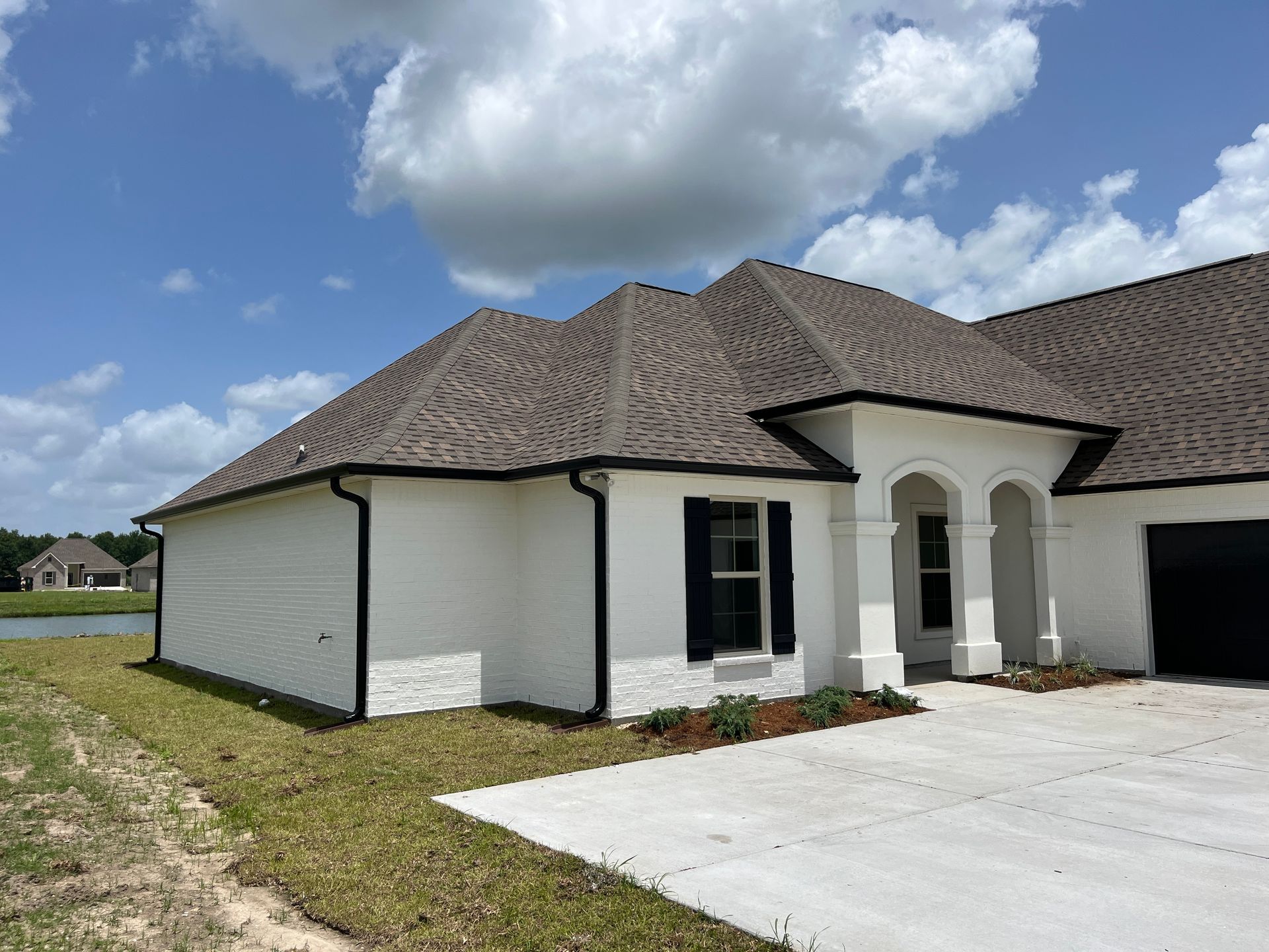 A single-story white brick house with black shutters and trim, under a blue sky with clouds, next to a concrete driveway.