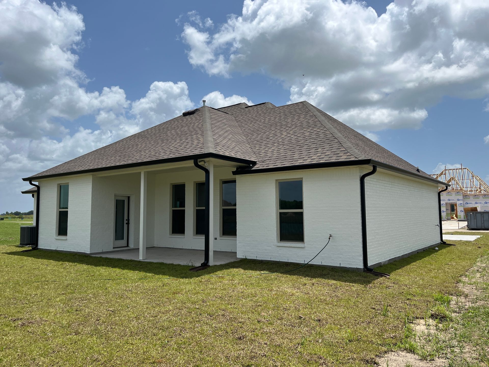 A white brick house with a dark roof and black gutters sits on a grassy lot under a blue sky with scattered clouds.