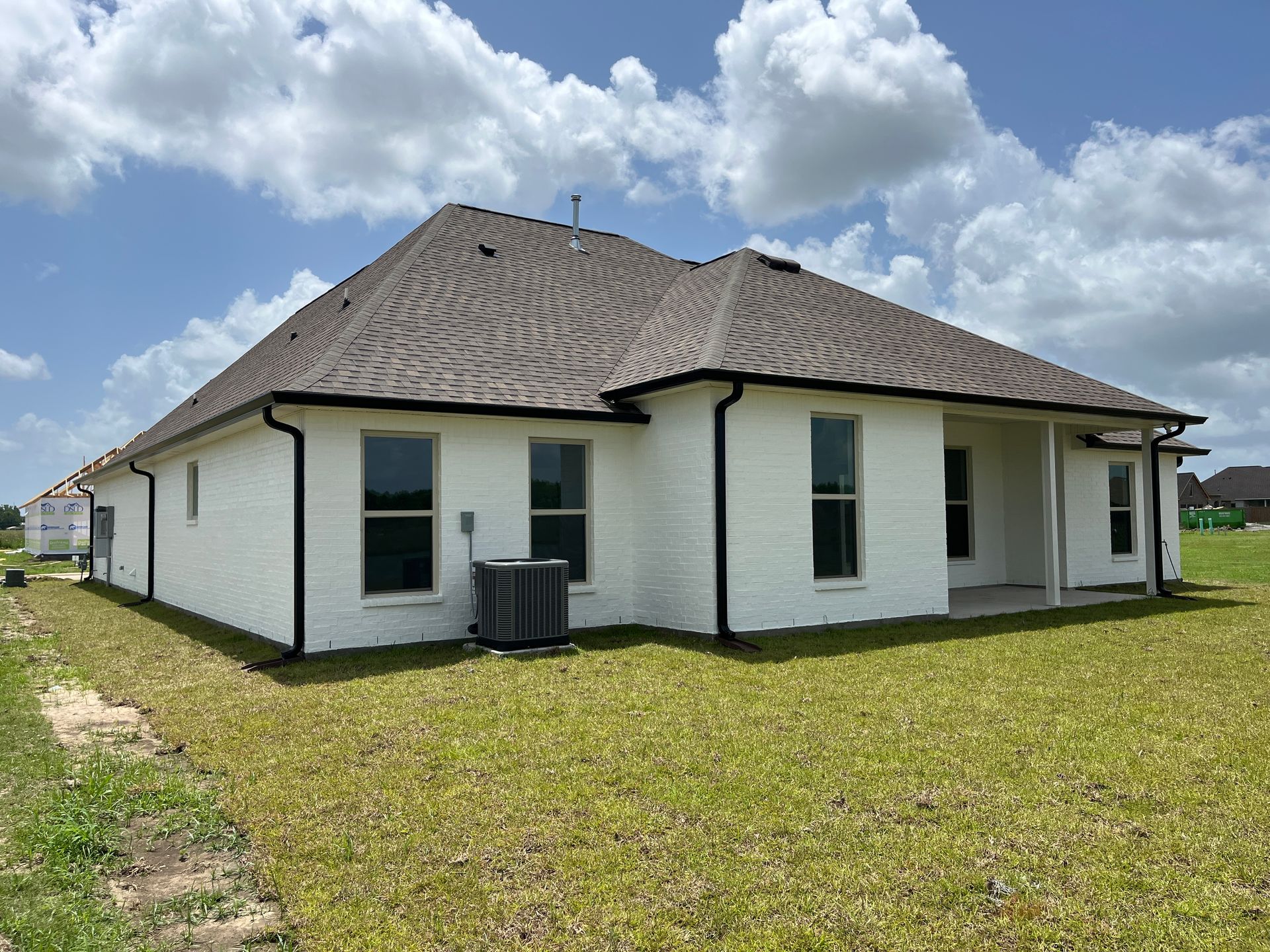 Back view of a white brick, single-story residential house with a dark shingled roof, set against a blue, cloudy sky.