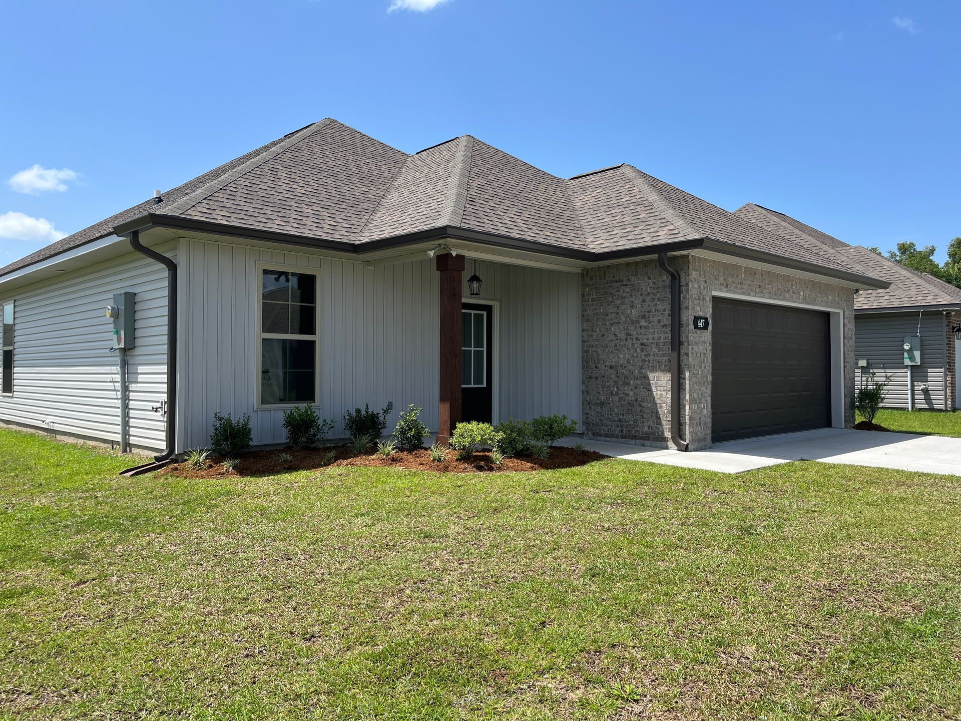A single-story suburban house with white vertical siding, a stone garage, and a grey shingled roof under a blue sky.