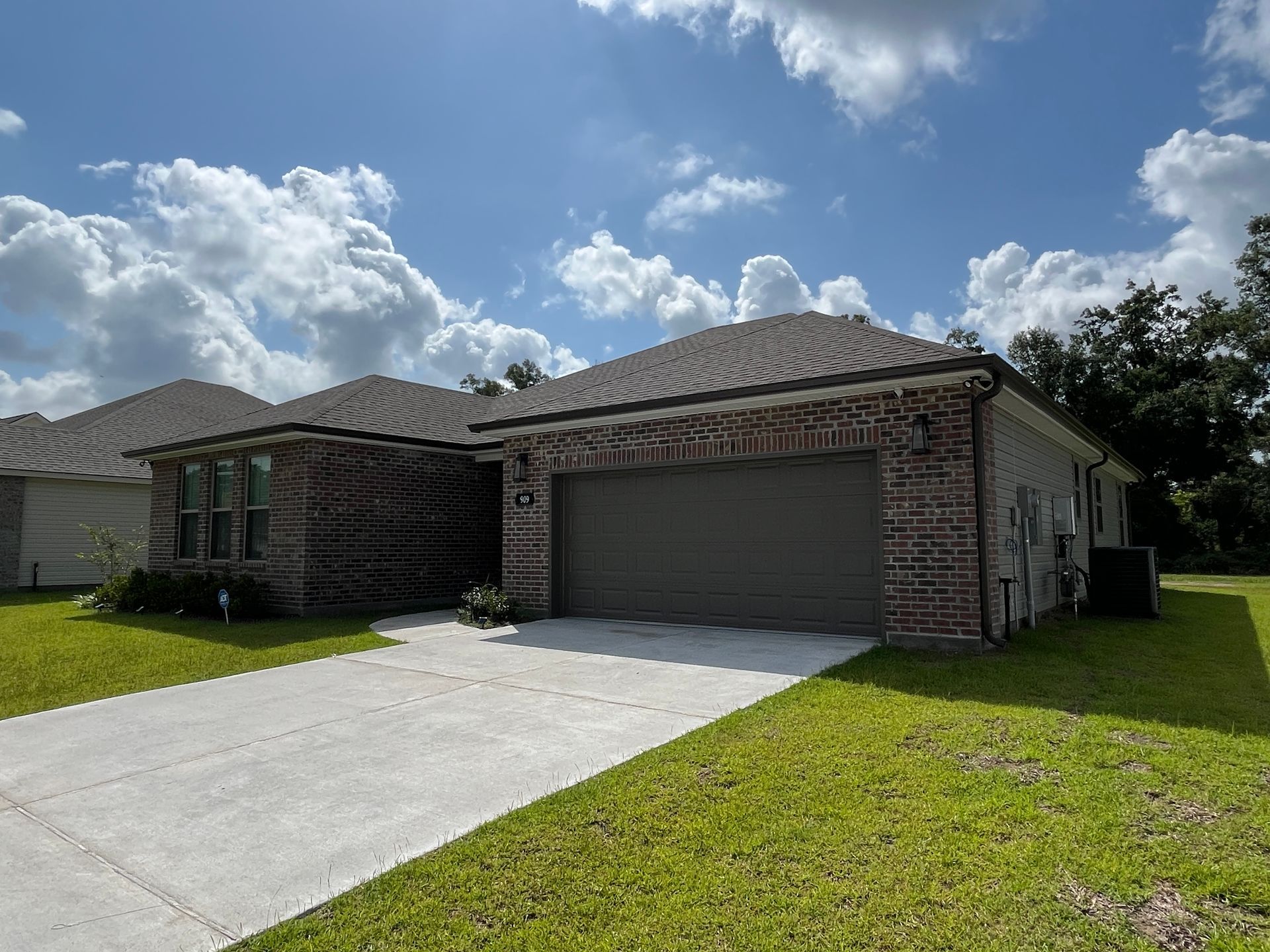 A single-story suburban house with a dark brick facade, gray tiled roof, and a two-car garage under a sunny blue sky.