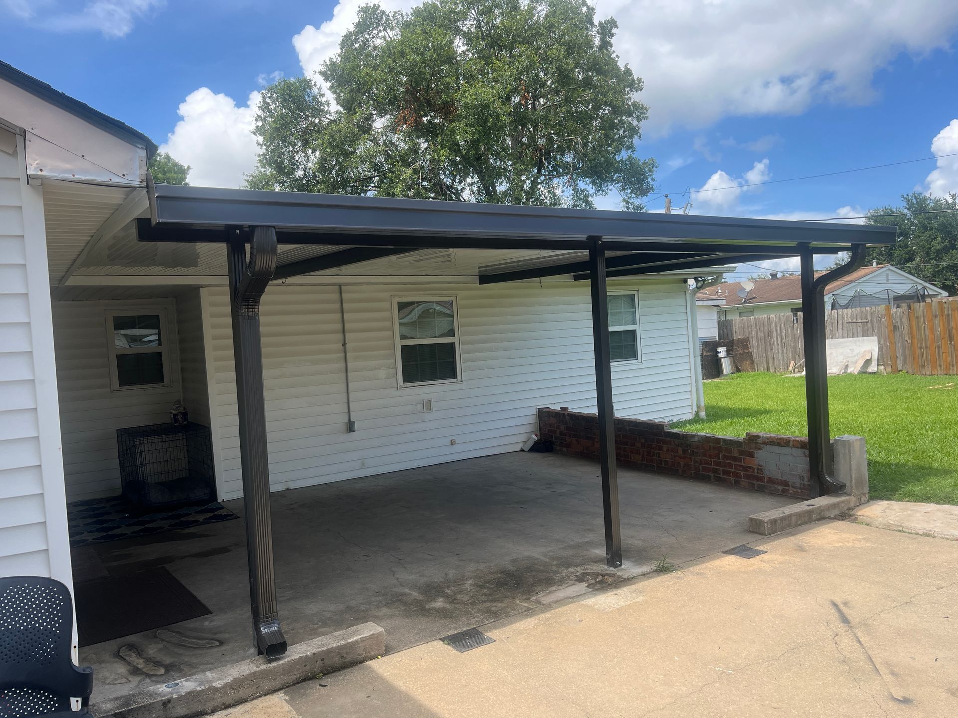 A covered patio area attached to a white house with a concrete slab floor, black metal posts, and a dark roof.