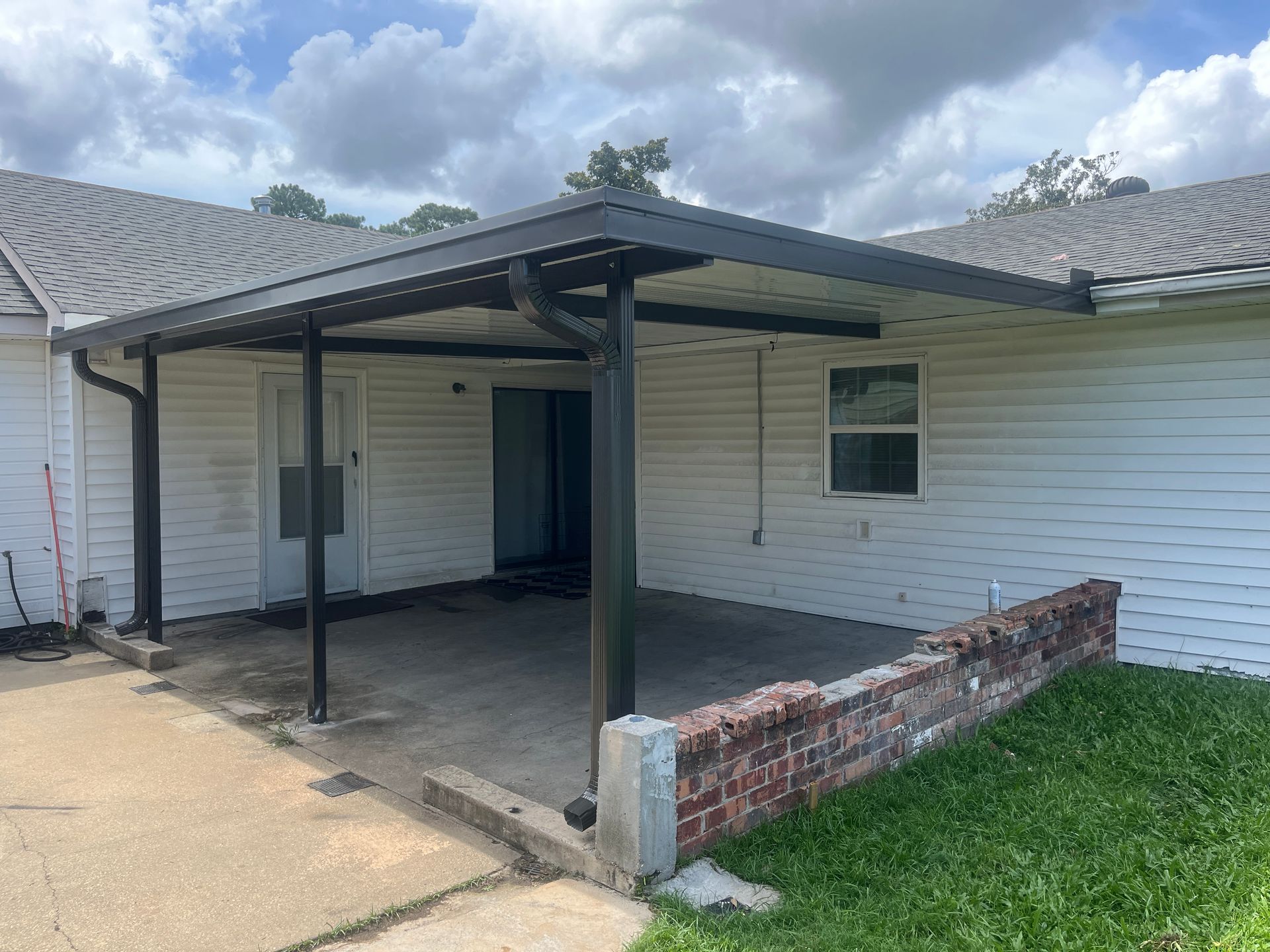 A white house exterior featuring a black covered patio area, a concrete driveway, and a low brick wall in the yard.