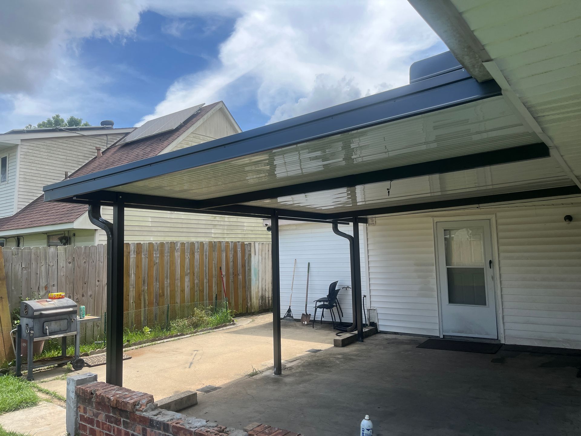 A black metal patio cover attached to a white-sided house with a concrete patio and a brick border.