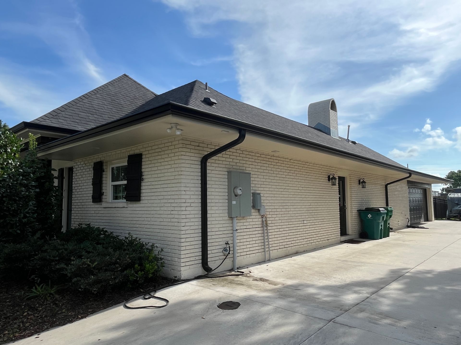 Exterior view of a cream-colored brick house with a dark shingle roof and black shutters under a blue sky.