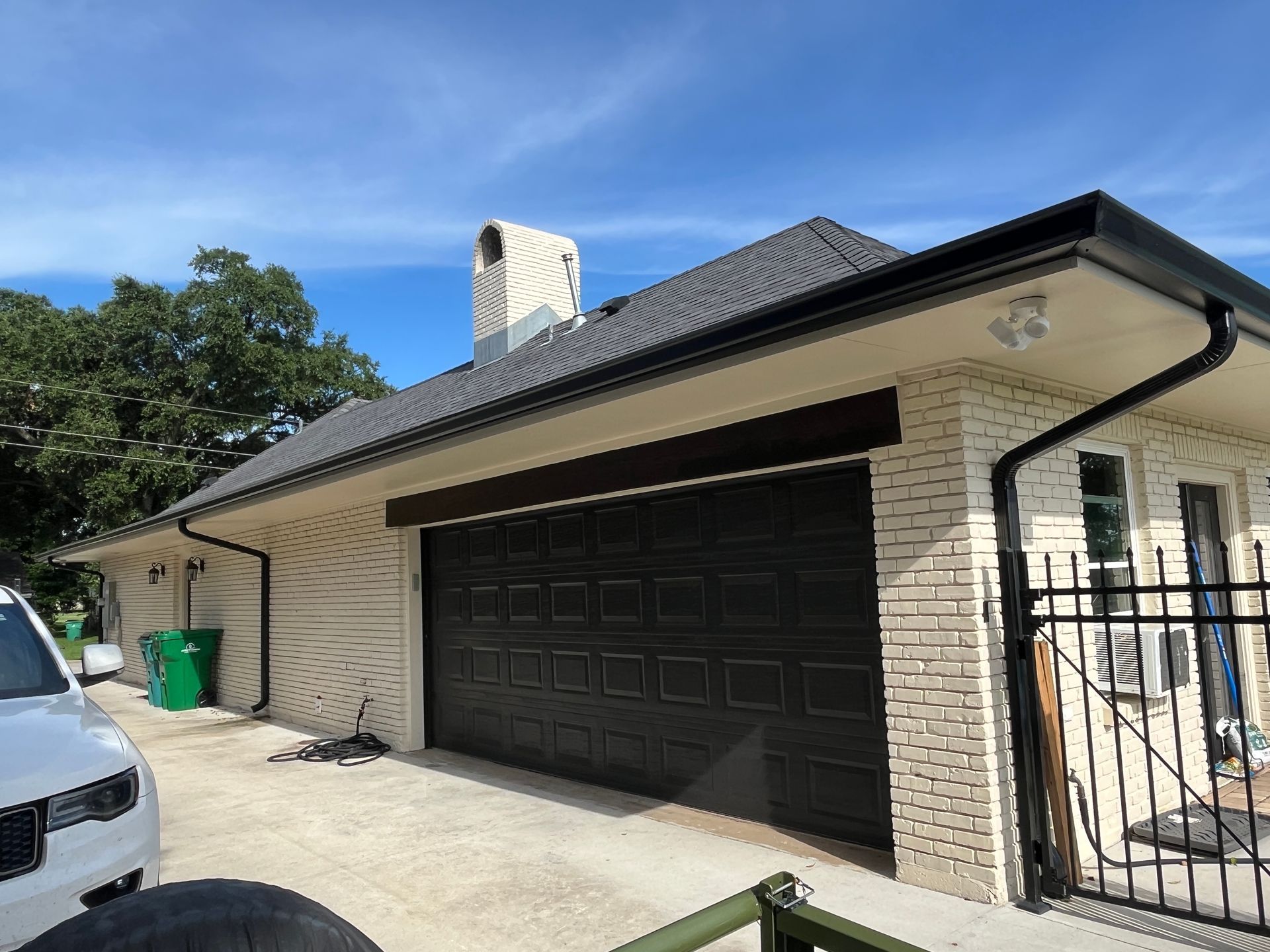 A brick house with a black garage door, a shingled roof, a chimney, and a parked car under a bright blue sky.