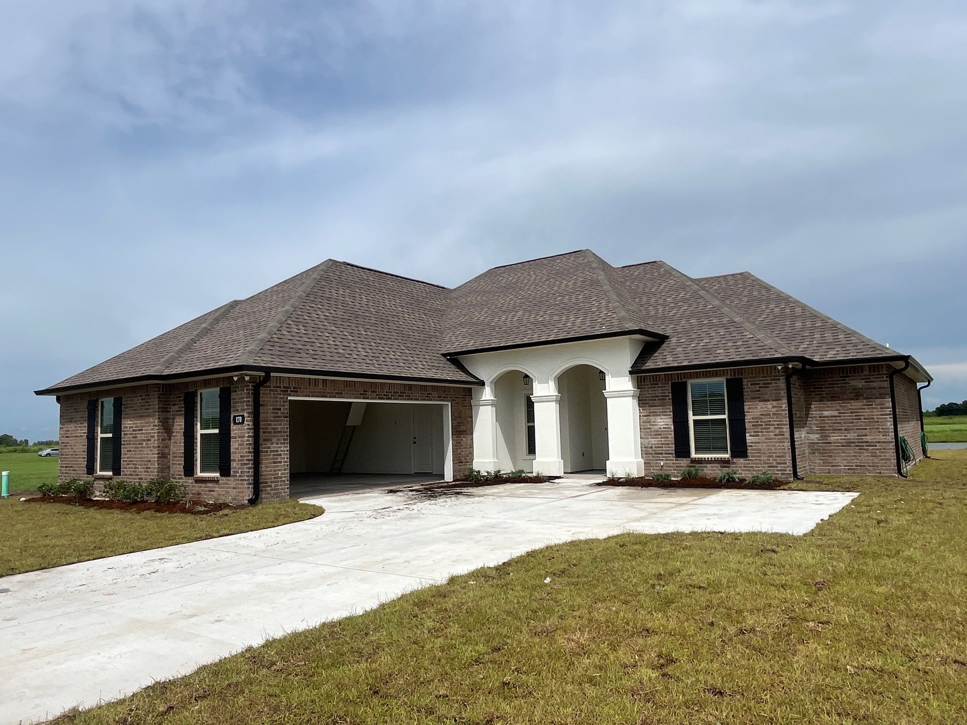A single-story brick house with a concrete driveway and arched entrance, set against a cloudy blue sky.