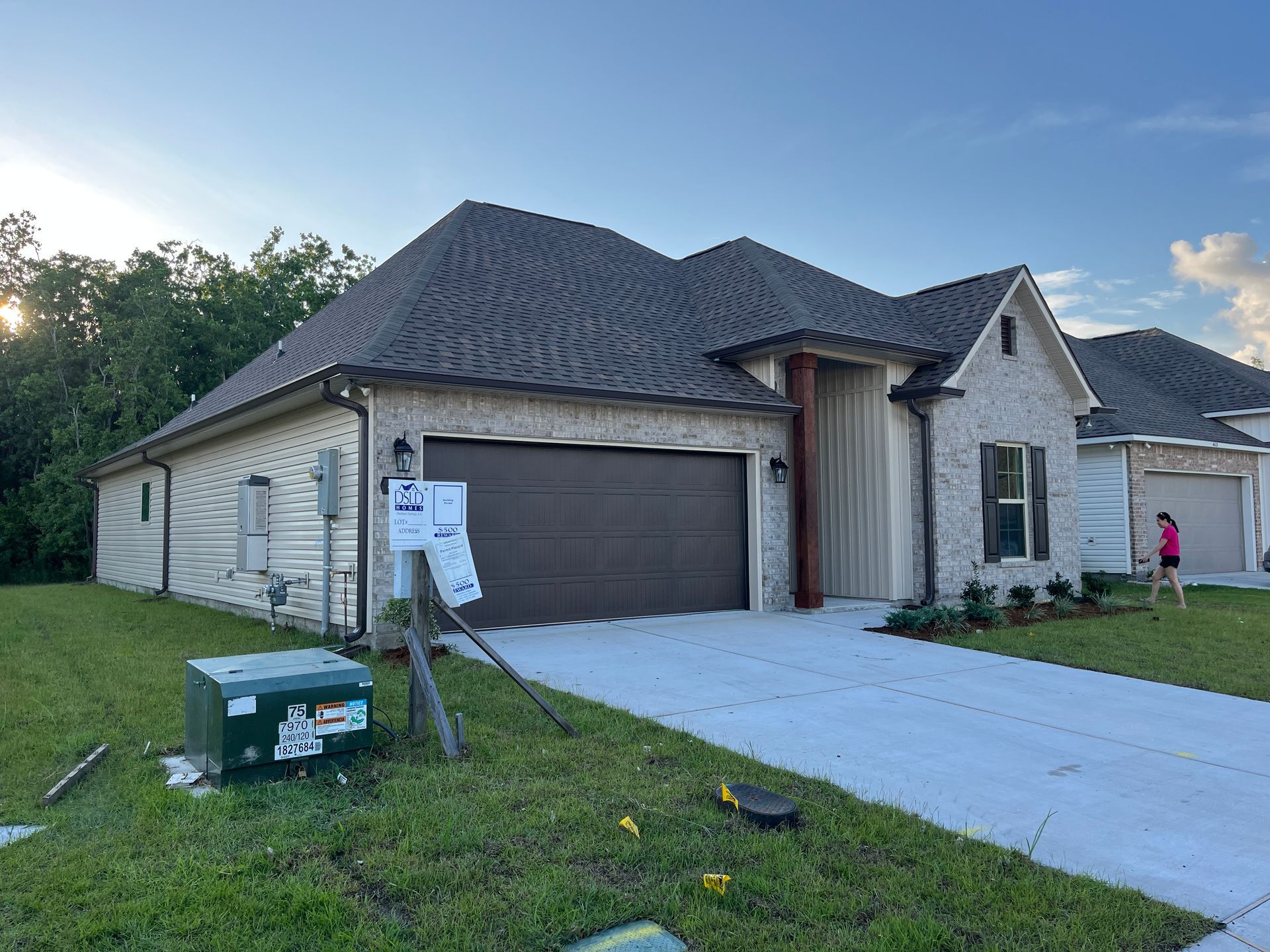 A tan brick house with a dark brown garage door, shingled roof, and green lawn under a clear blue sky.