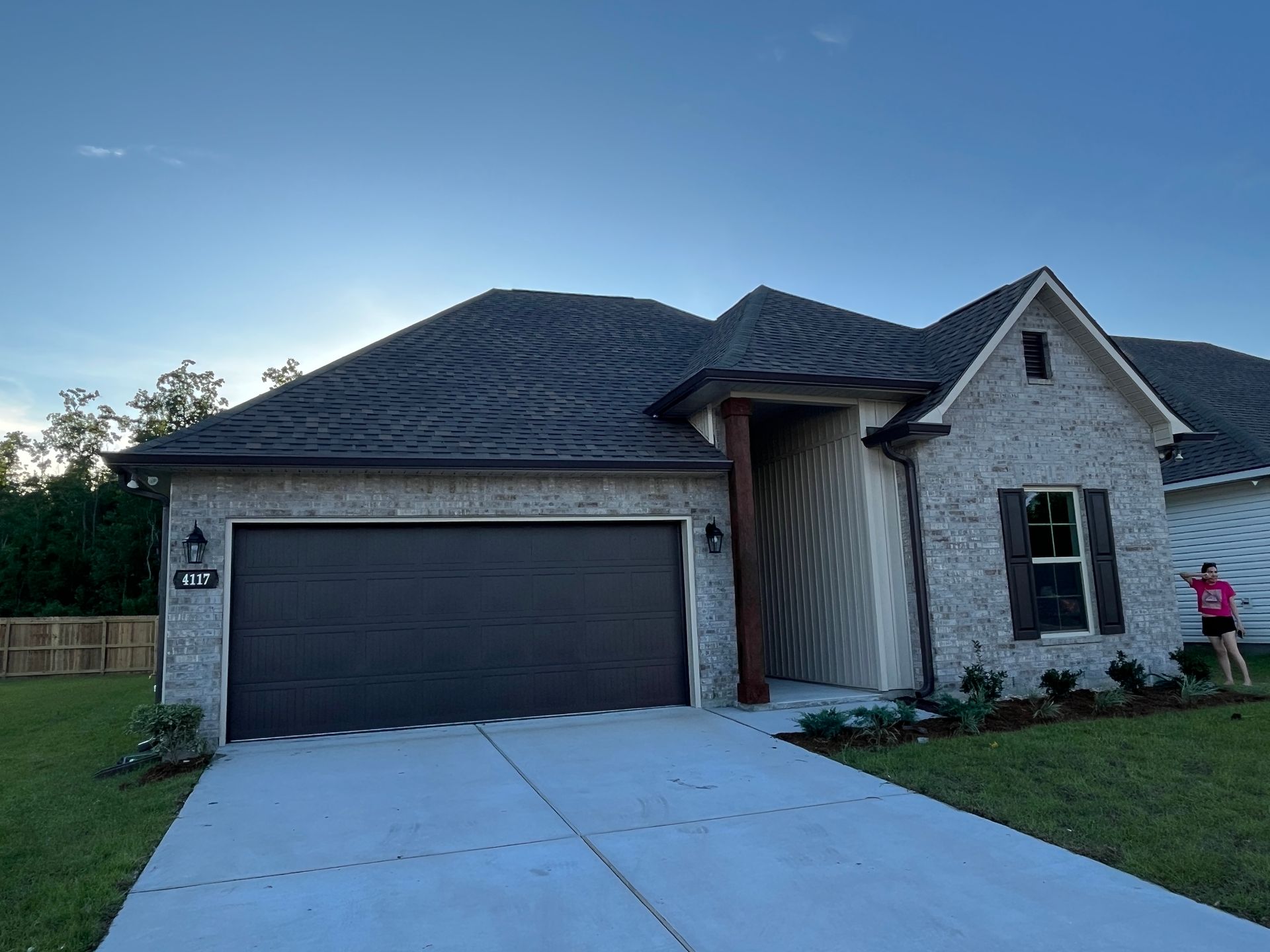 A modern, single-story house with gray brick, a dark garage door, a peaked roof, and a person walking on the front lawn.