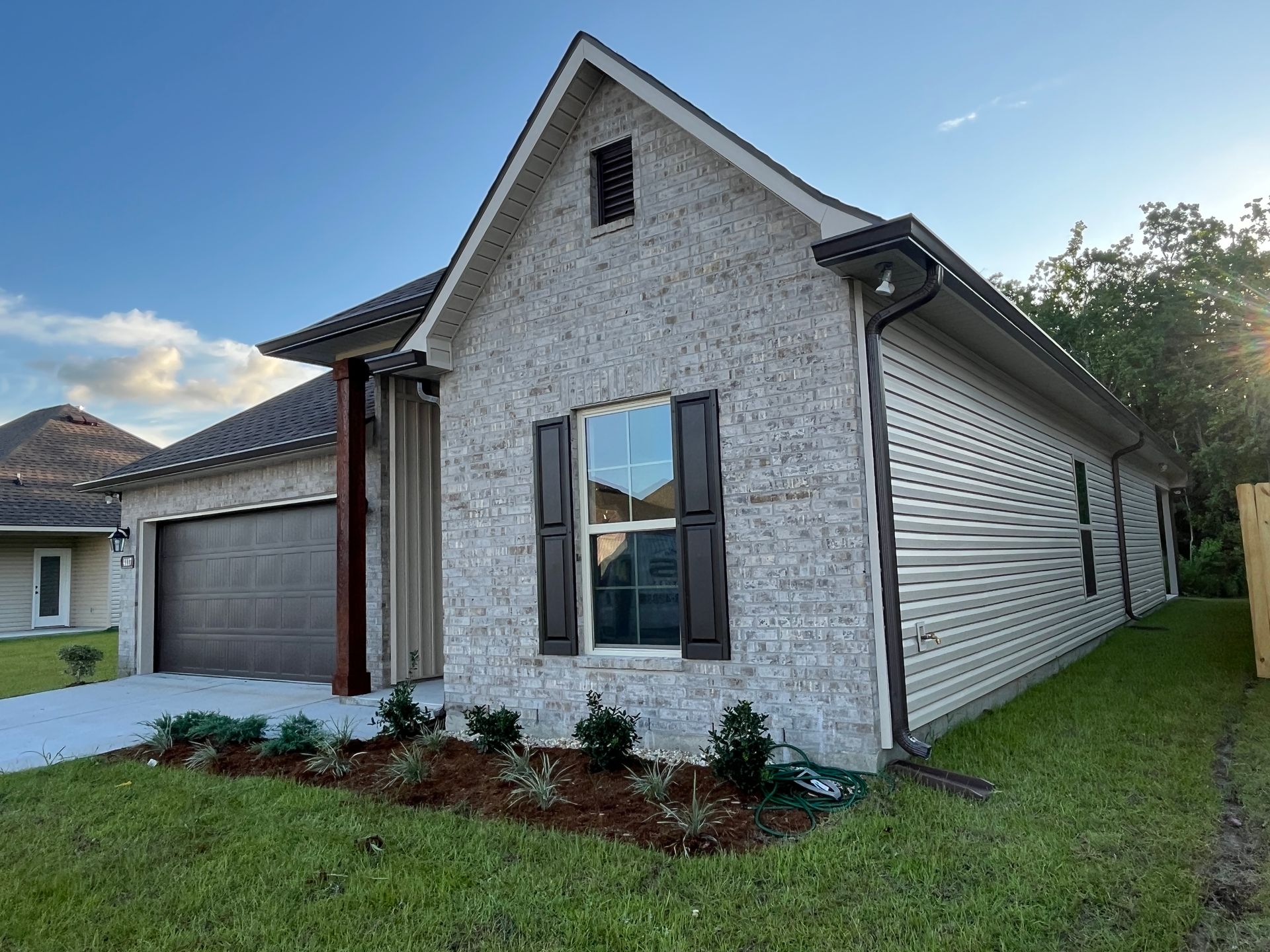 A modern house with a gray brick front, dark brown garage door, and light-colored siding on a sunny, grassy lawn.