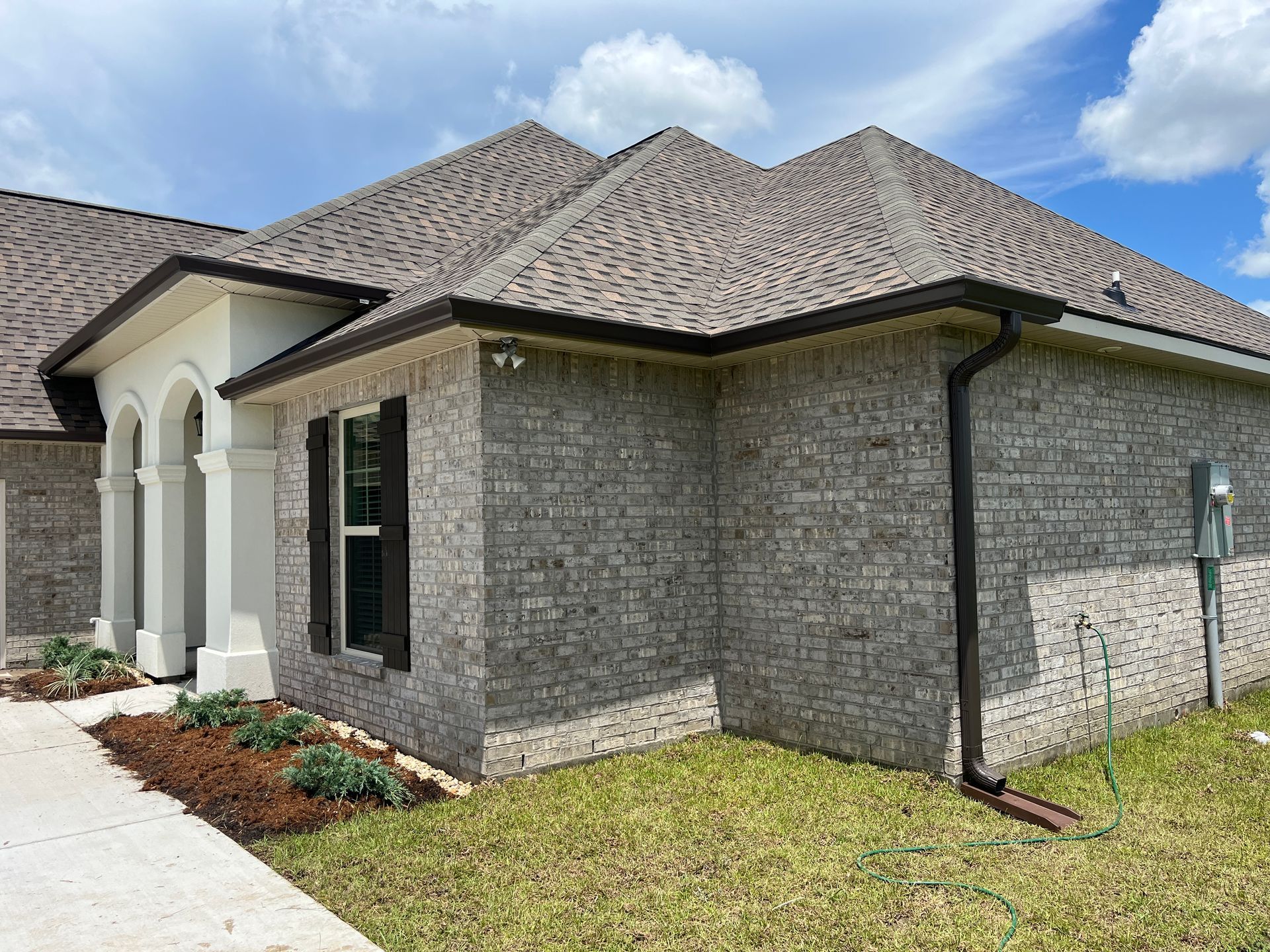 Exterior view of a modern one-story brick home with a gray shingled roof, arched entryway, and dark shutters.