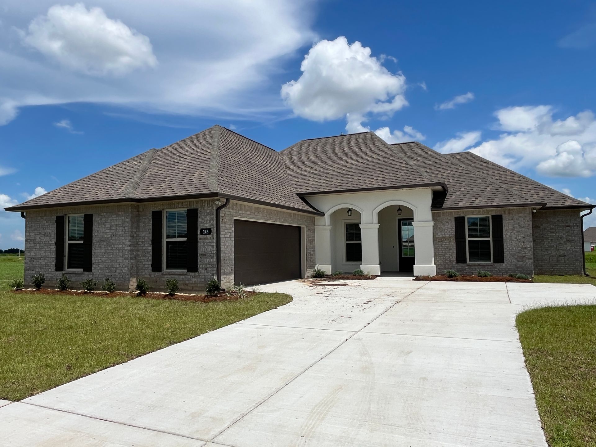 A one-story brick house with a concrete driveway, brown roof, and white arched entryway under a bright blue sky.