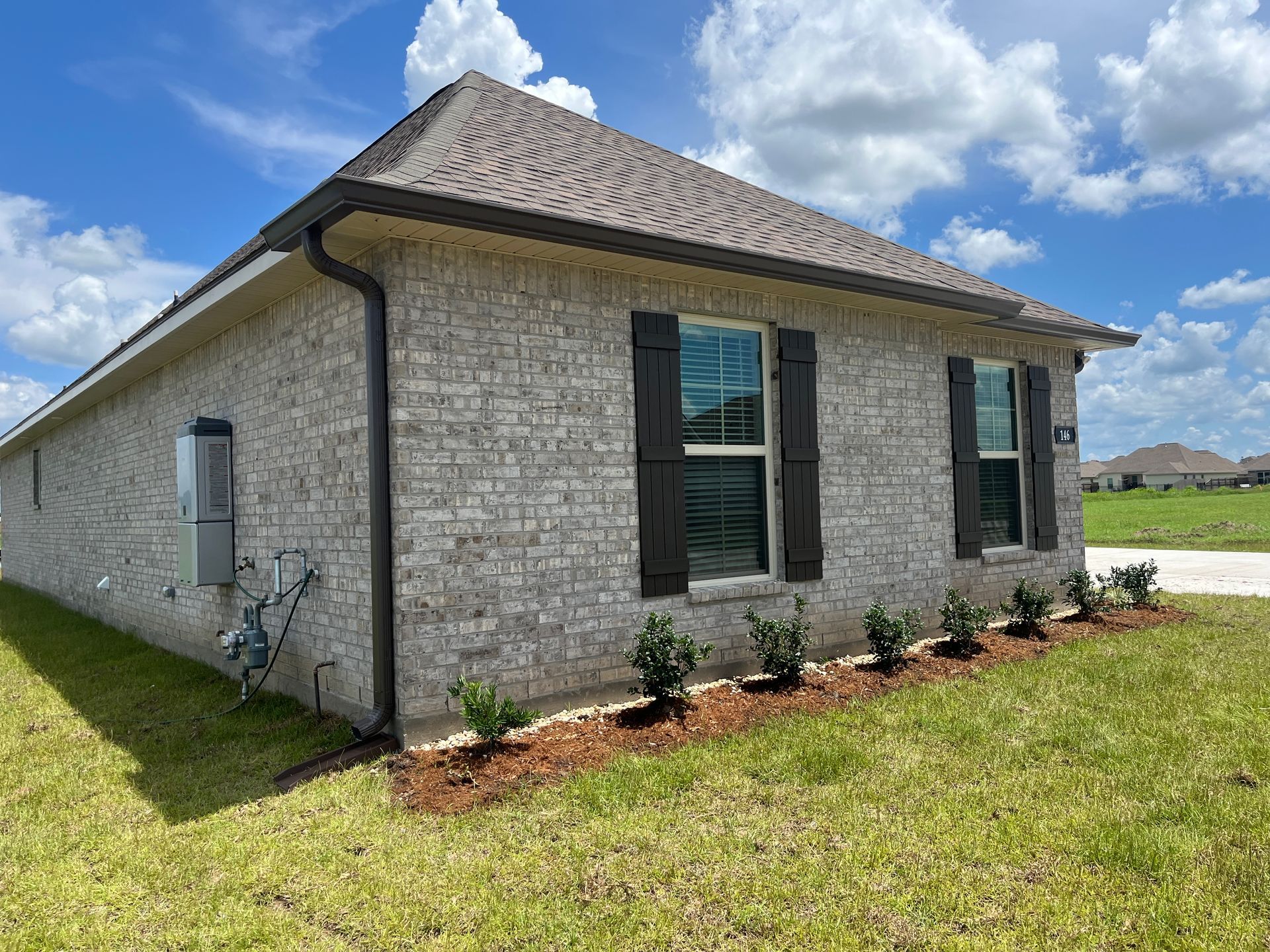 Side view of a light-colored brick residential house with dark shutters and a small garden bed against a blue sky.