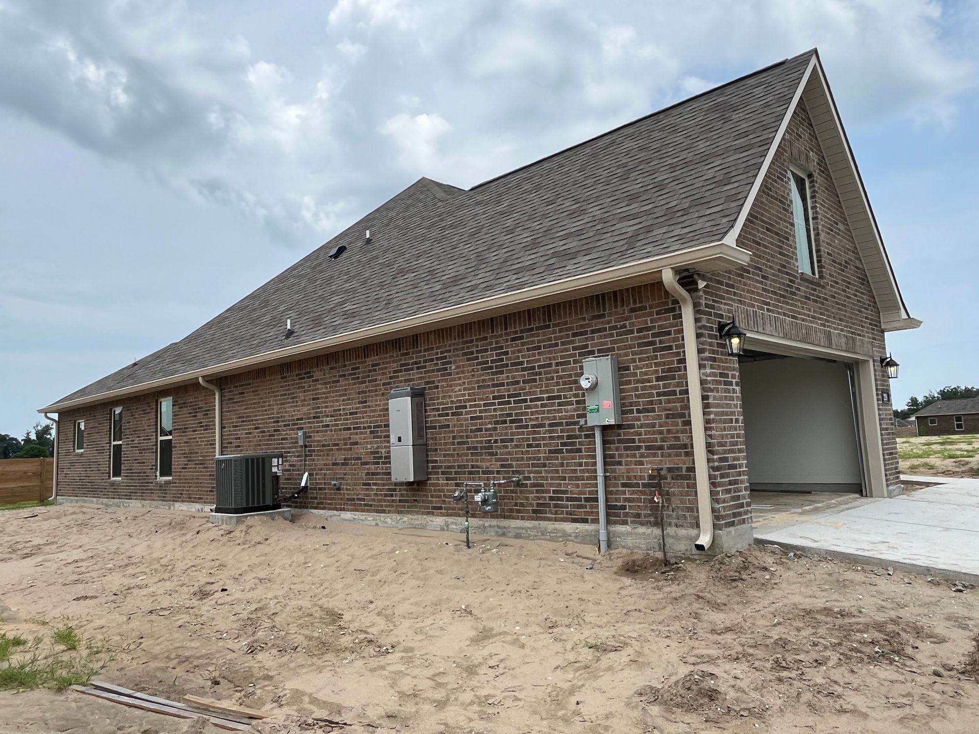 A side view of a new brown brick house with a garage, utility meters, and an air conditioning unit on a dirt lot.