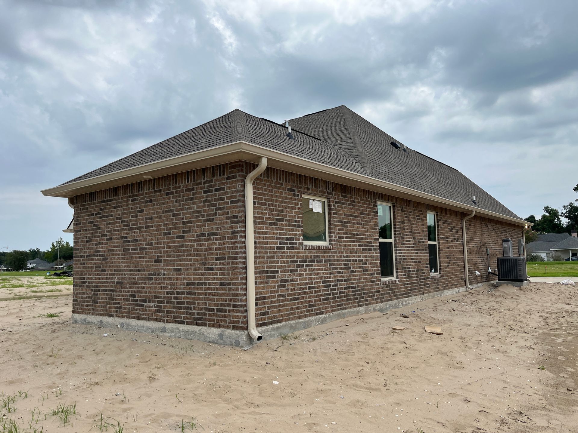 A side view of a new brown brick single-story house with tan trim under a cloudy sky on an undeveloped sandy lot.