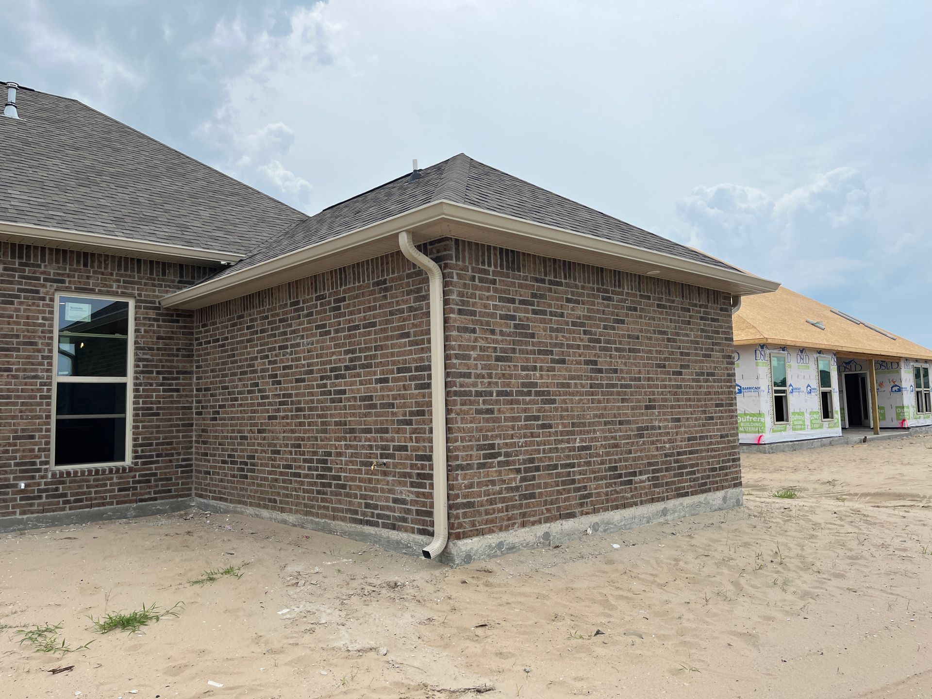 Exterior of a brick house under construction with a tan downspout, adjacent to another home with wood framing.