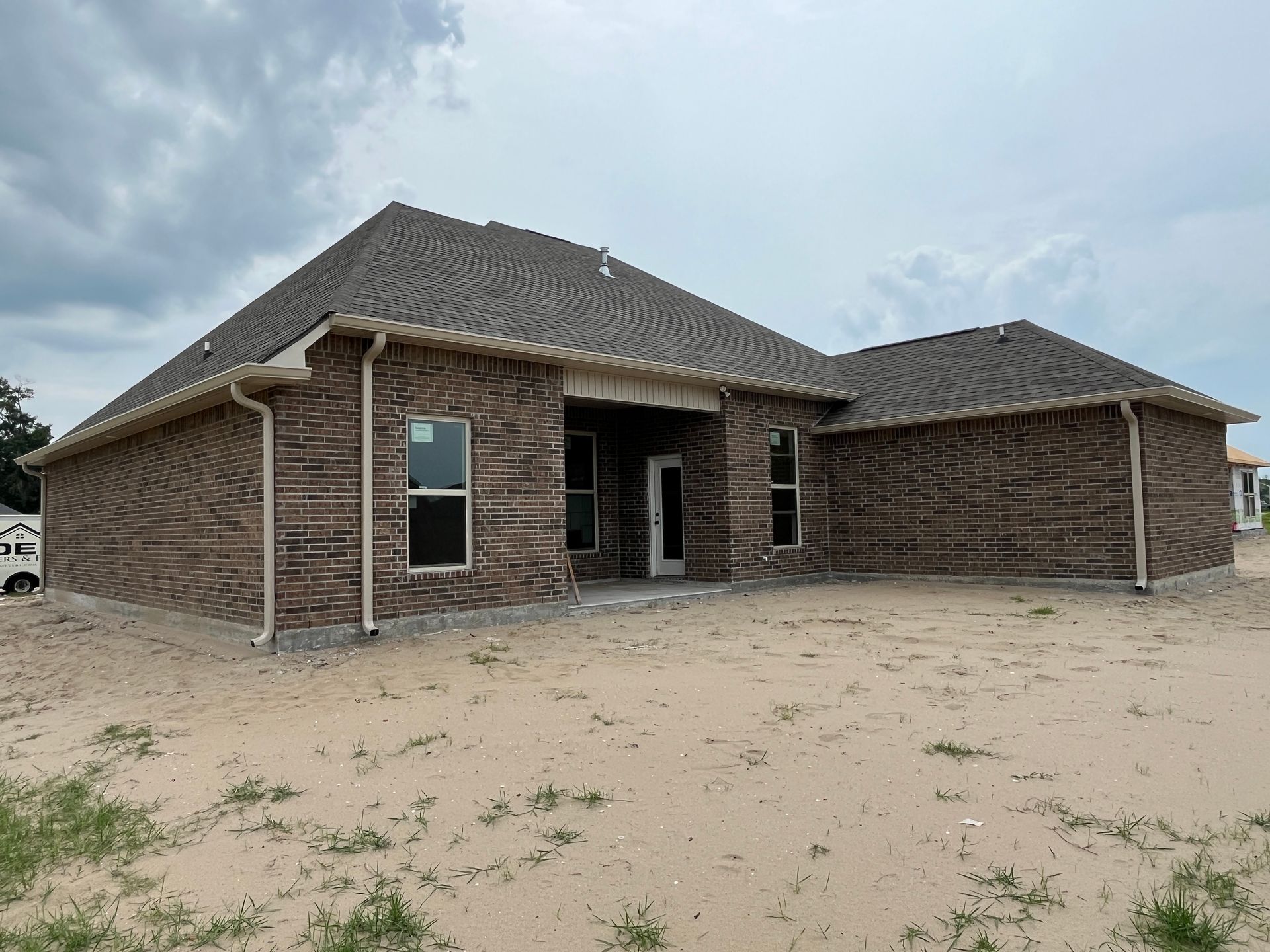 A newly constructed, single-story brick house with a dark shingled roof under a cloudy sky on a sandy, undeveloped lot.