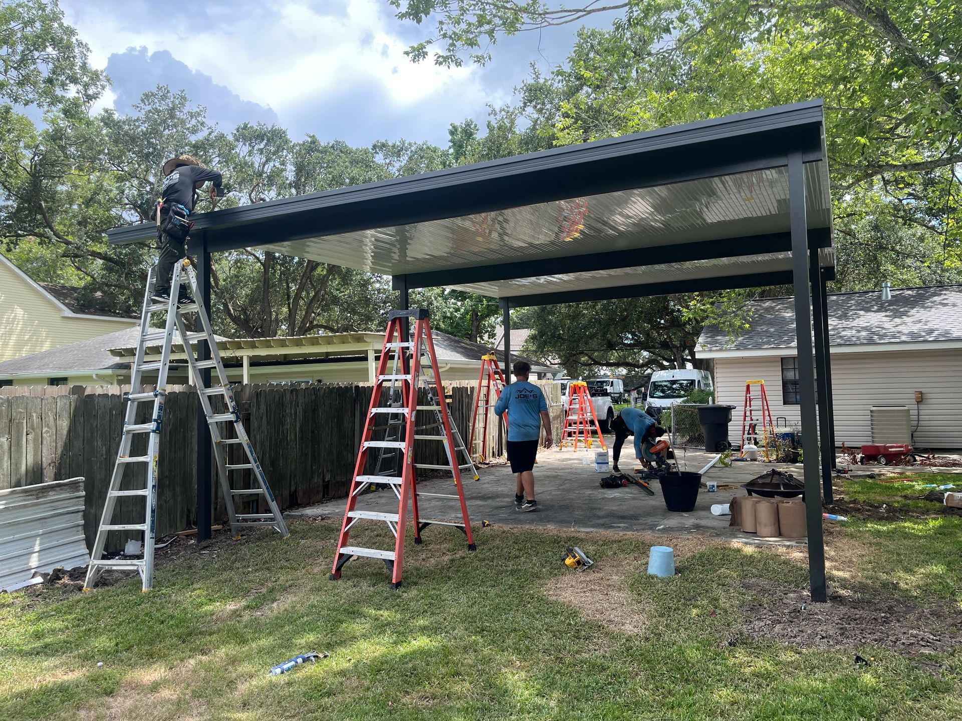 Workers assemble a black metal patio cover in a backyard, using ladders on a paved area near a house and trees.