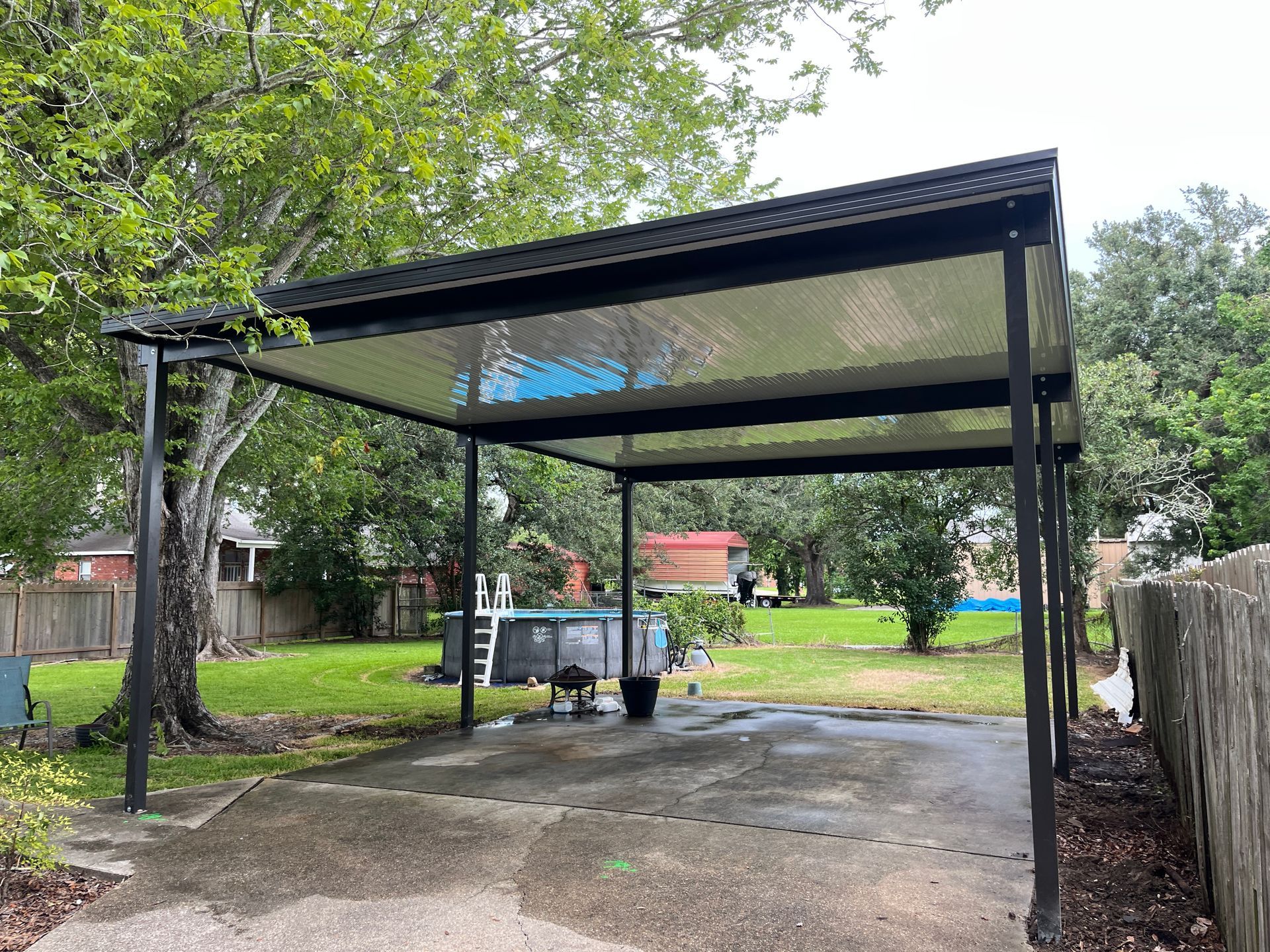 A black, modern metal carport with a translucent roof stands over a concrete driveway in a grassy backyard.