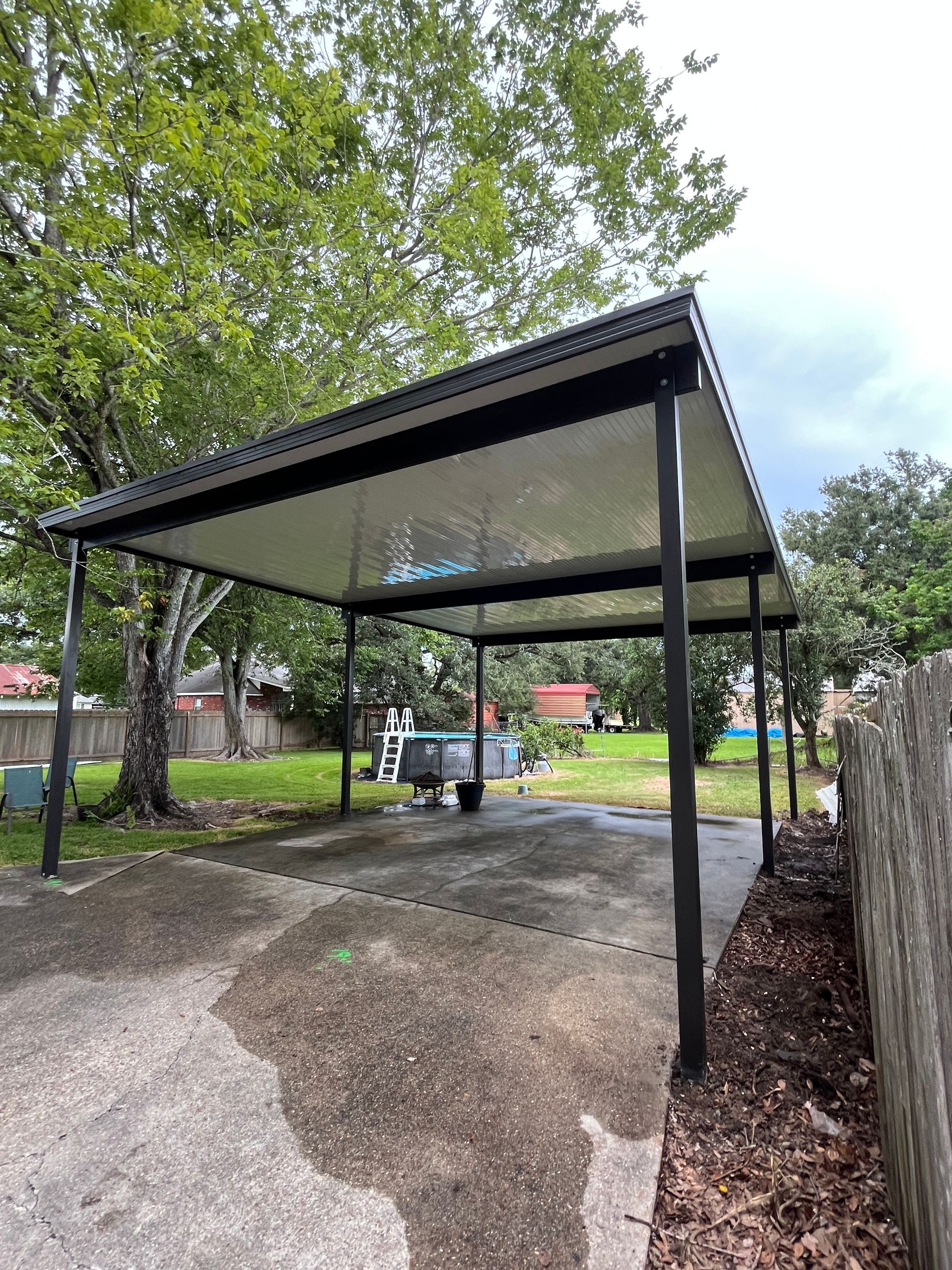 A dark metal carport with a flat roof stands over a concrete driveway in a backyard with trees and a wooden fence.