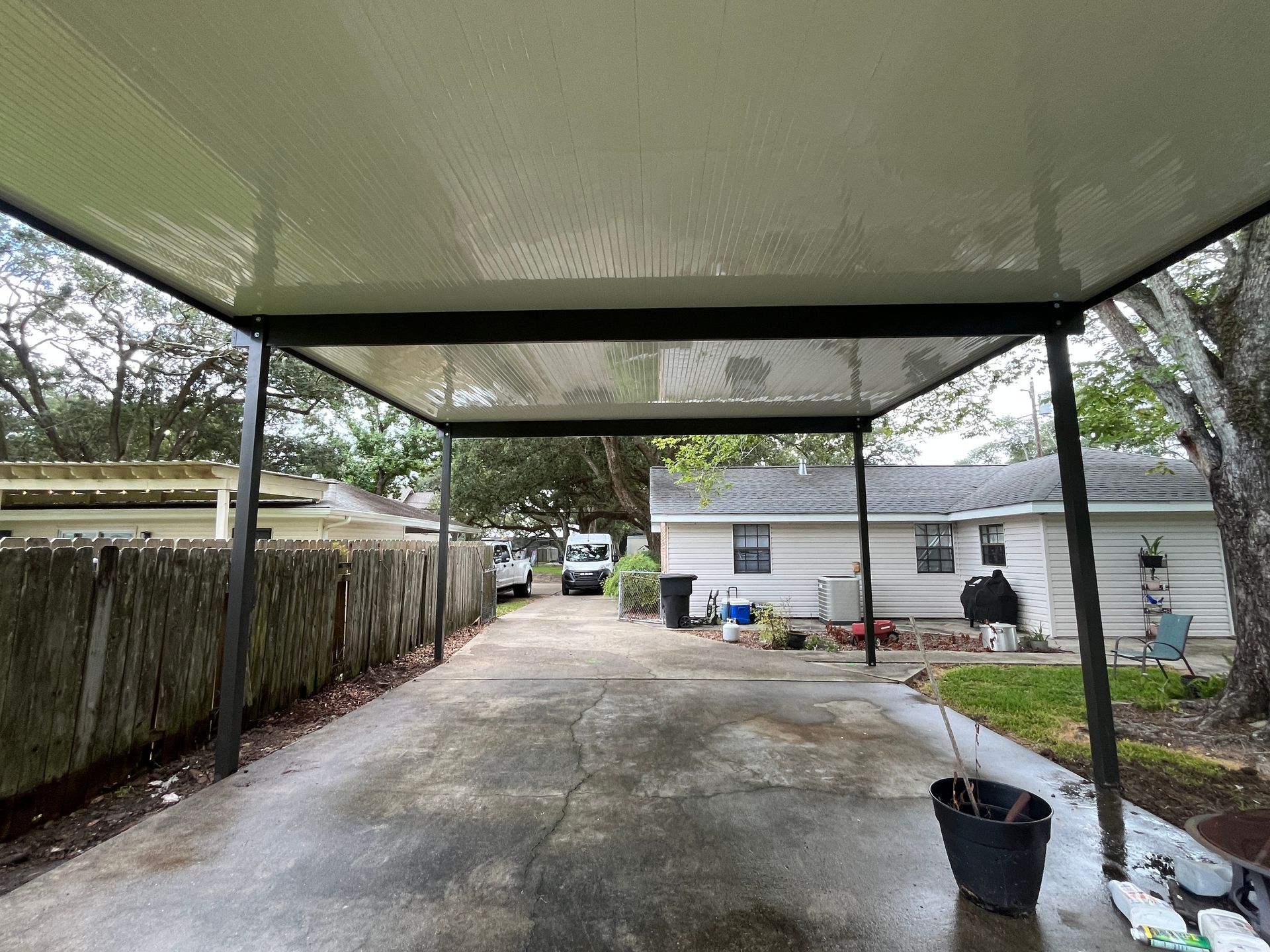 A metal carport covers a concrete driveway, looking toward a residential house, trees, and a parked utility vehicle.