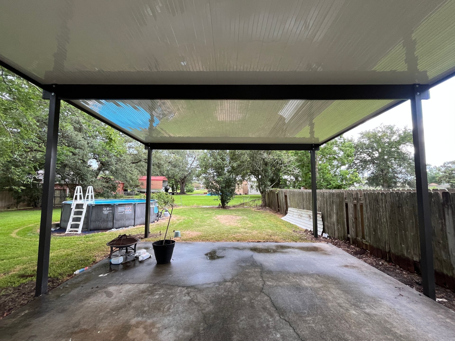 A view from under a black-framed patio cover looking out toward a backyard with a swimming pool and a stone wall.