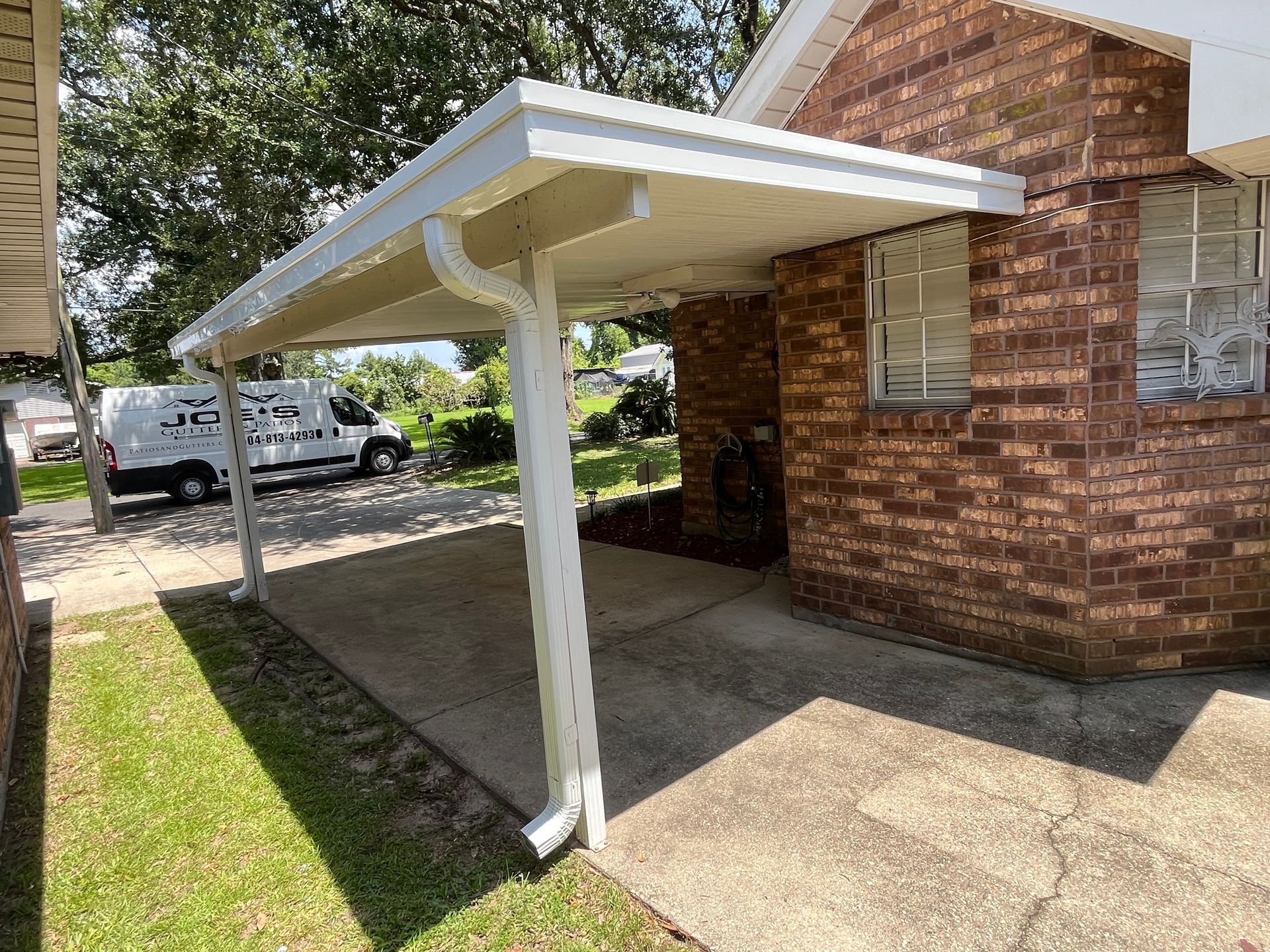 A brick house exterior with a white metal carport covering a concrete driveway on a sunny day.
