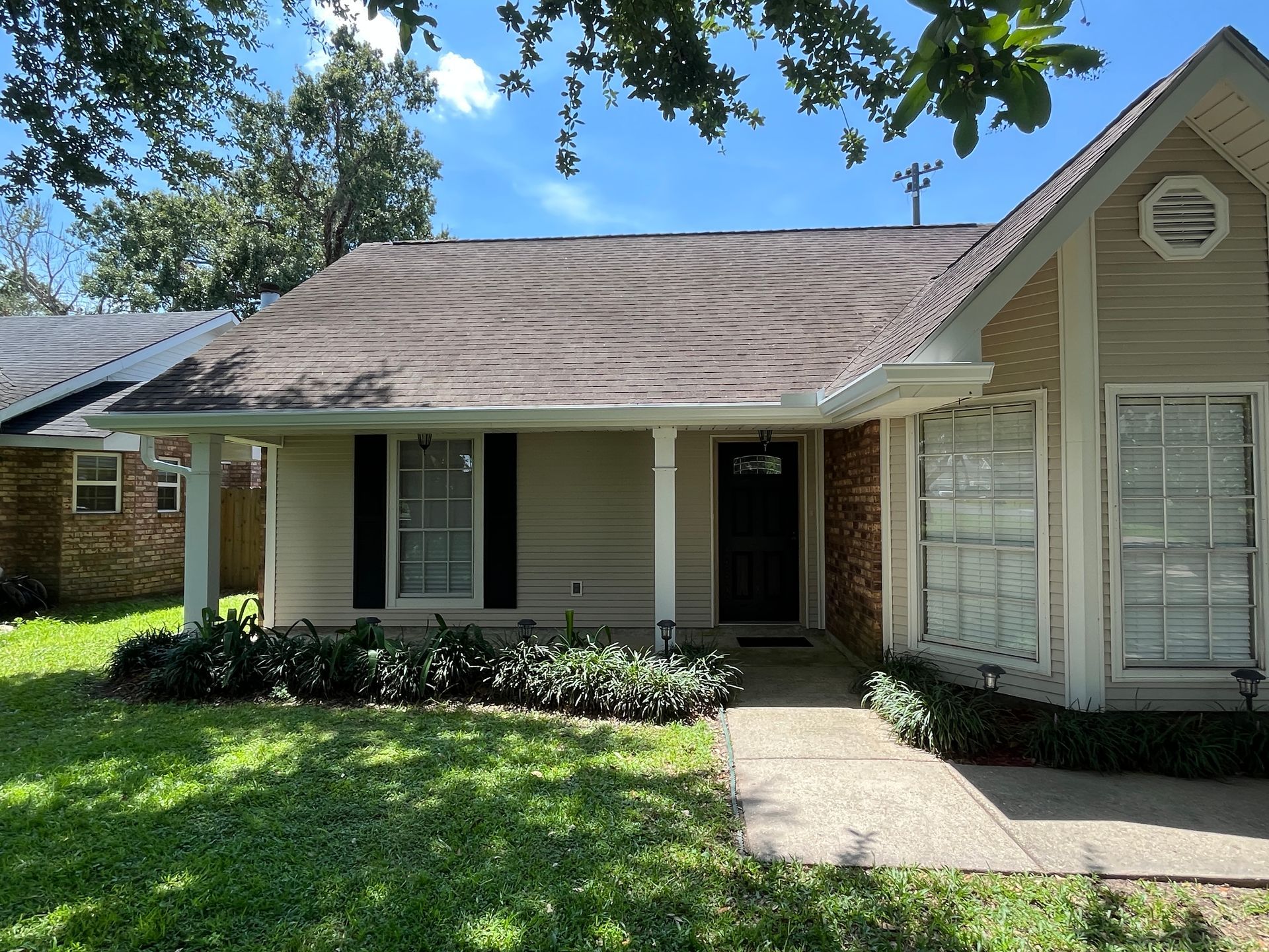 A one-story tan house with a dark roof, black shutters, a covered porch, and a walkway surrounded by green landscaping.