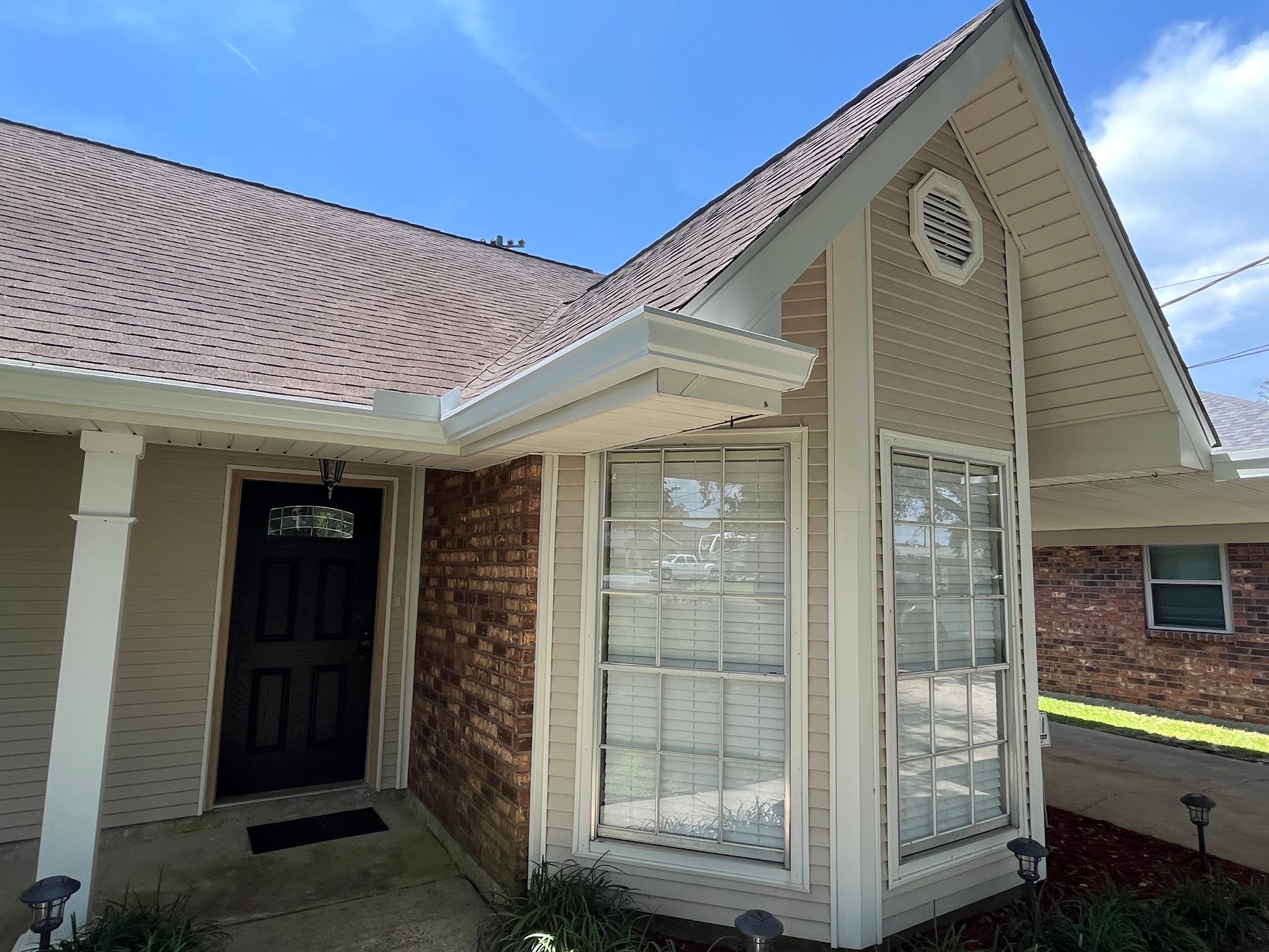 A beige house exterior features a brick wall, a black front door, and a gabled roof section above a large bay window.