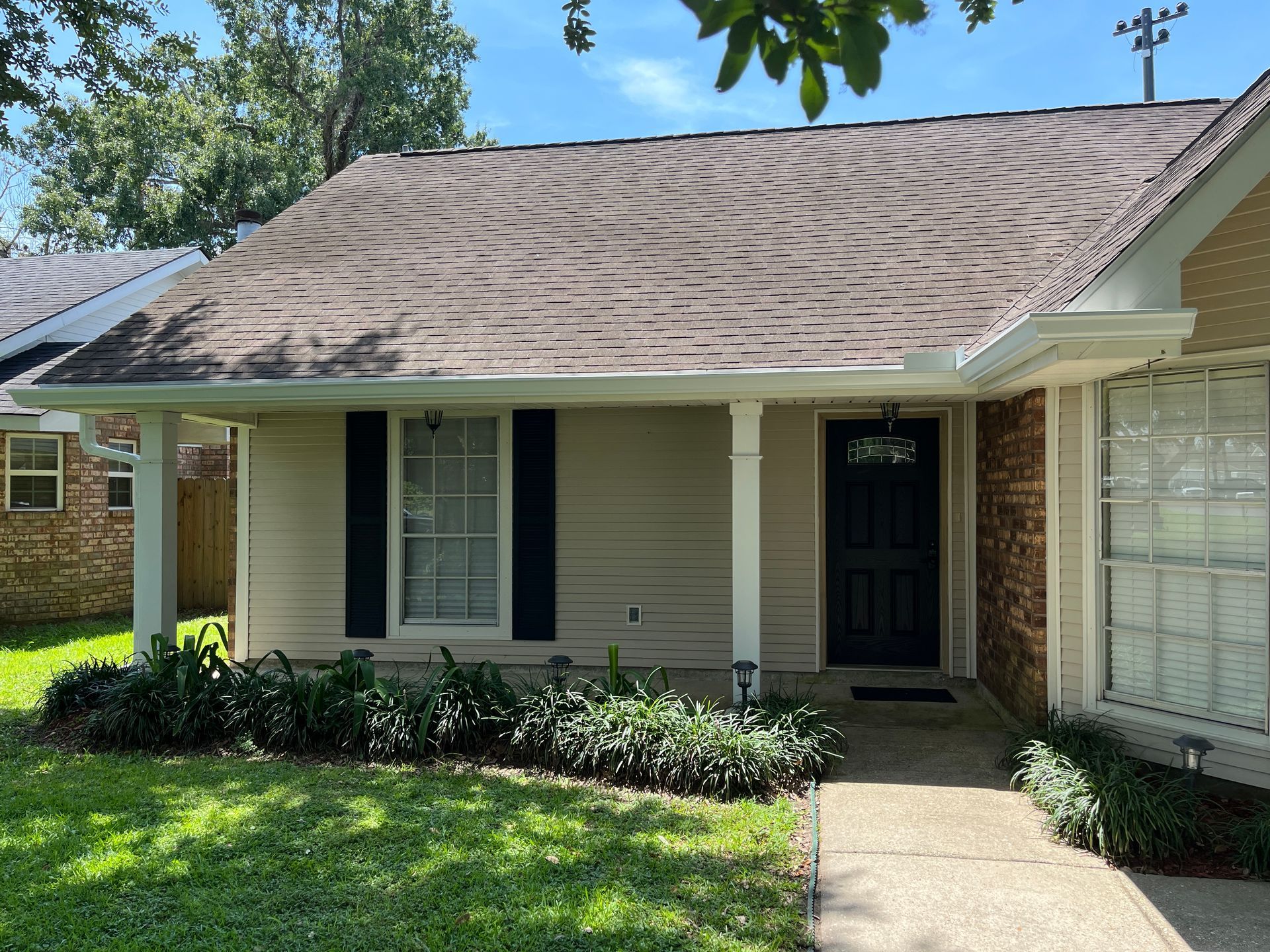 A single-story, tan-sided house with a dark roof, black shutters, a dark front door, and a landscaped front yard.