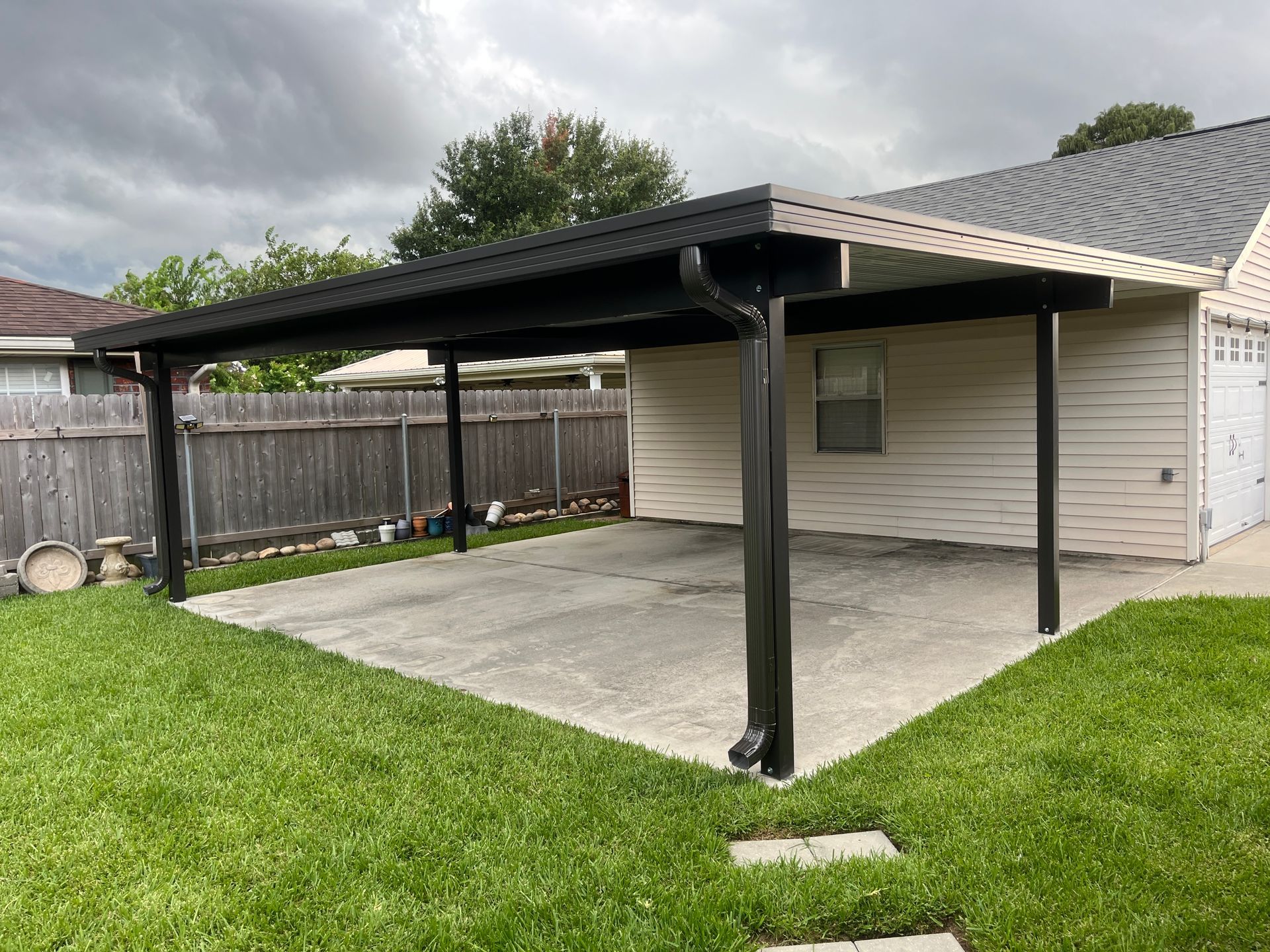 A black metal carport structure stands over a concrete driveway next to a light-colored house with a gray roof.