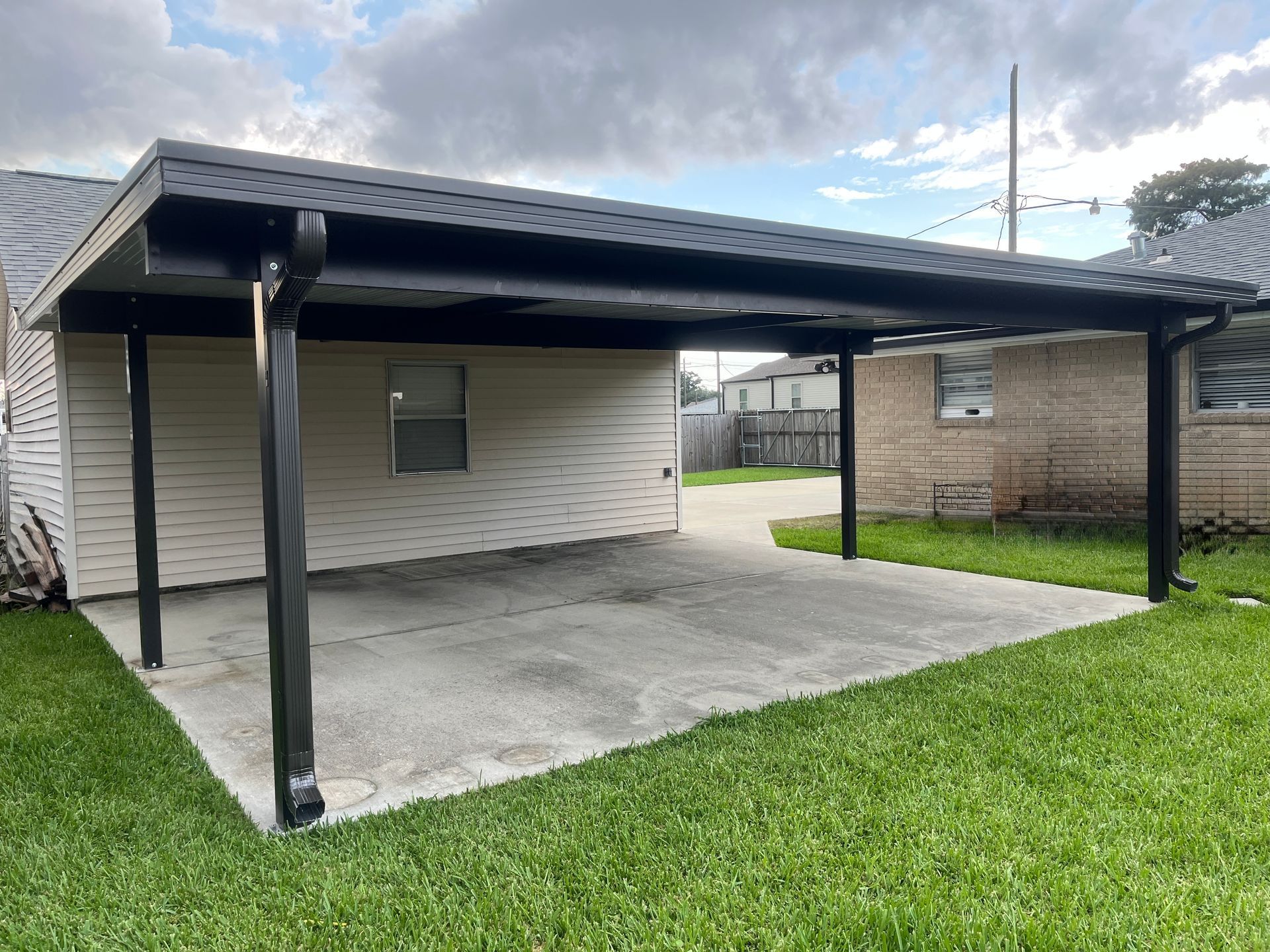 A black metal carport covering a concrete patio area, attached to a house with beige vinyl siding and green grass.