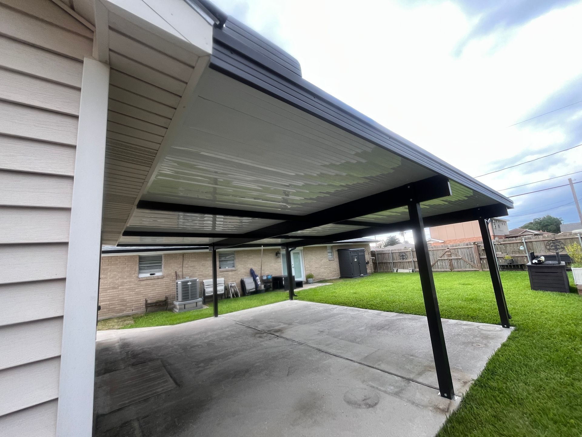 A black metal patio cover attached to a house, extending over a concrete driveway area with a grassy yard in the background.