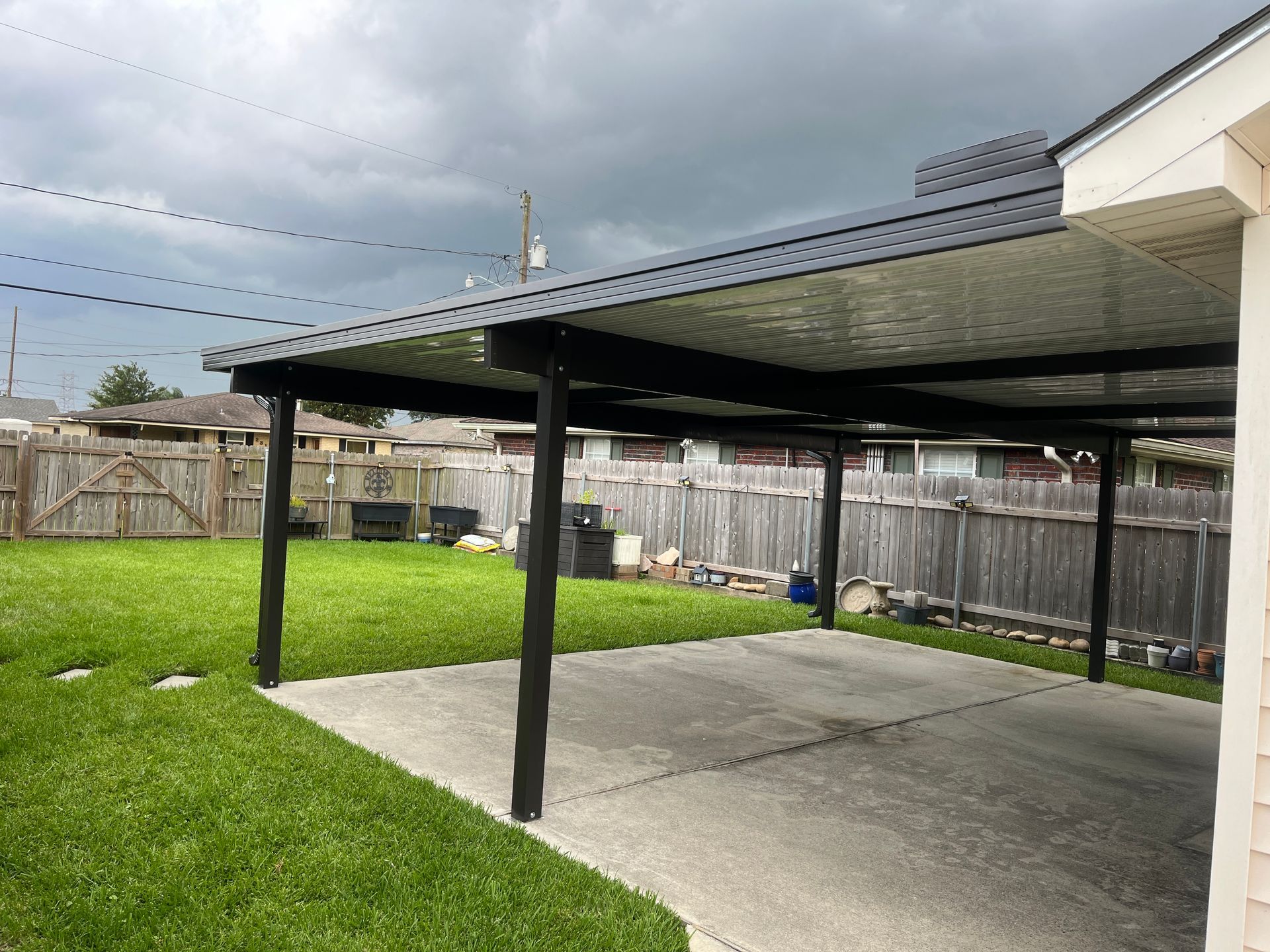 A black covered patio structure stands over a concrete slab in a grassy backyard under a cloudy sky.