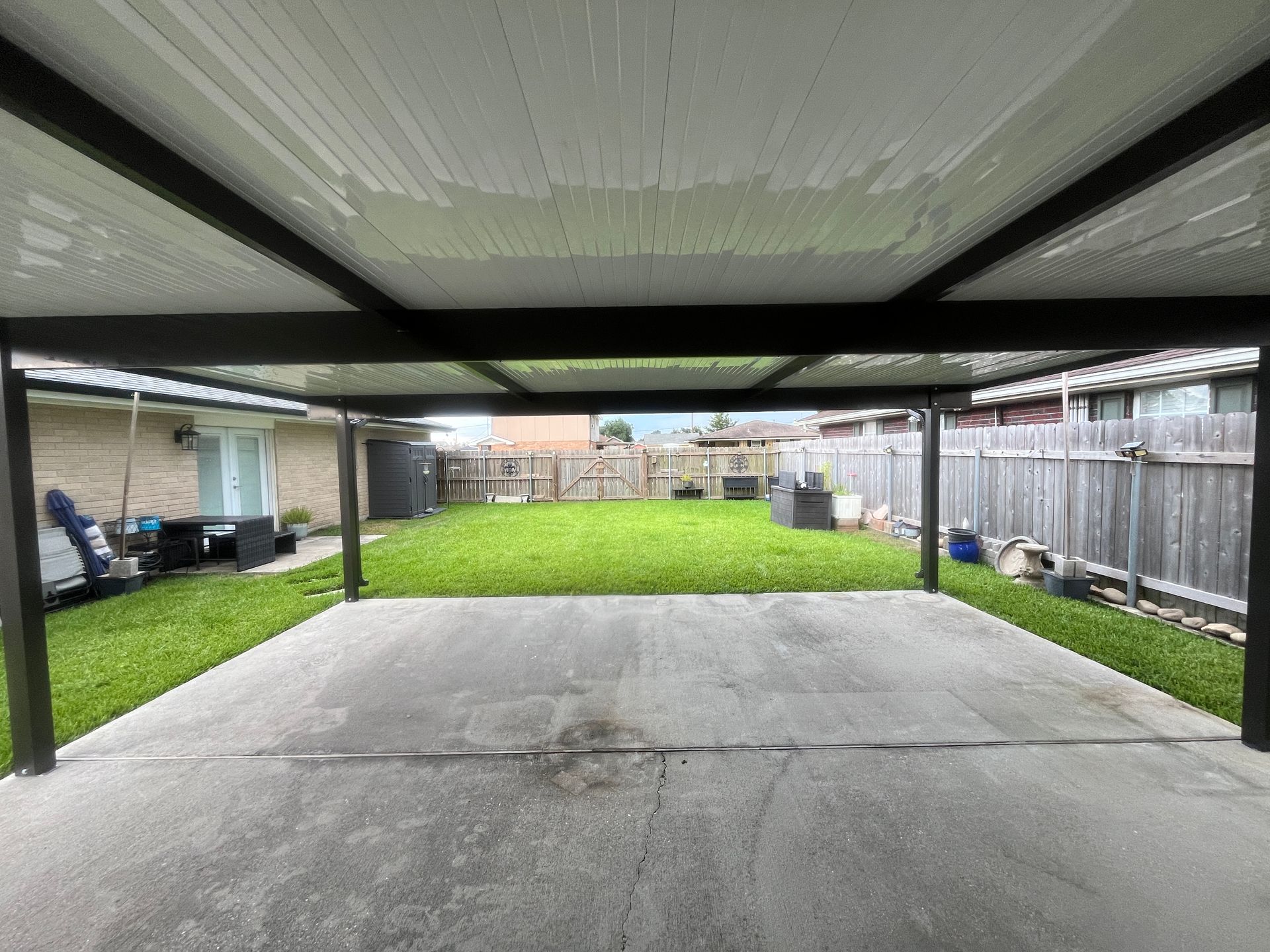 A concrete carport with a metal roof looking out onto a grassy backyard with a wooden fence.