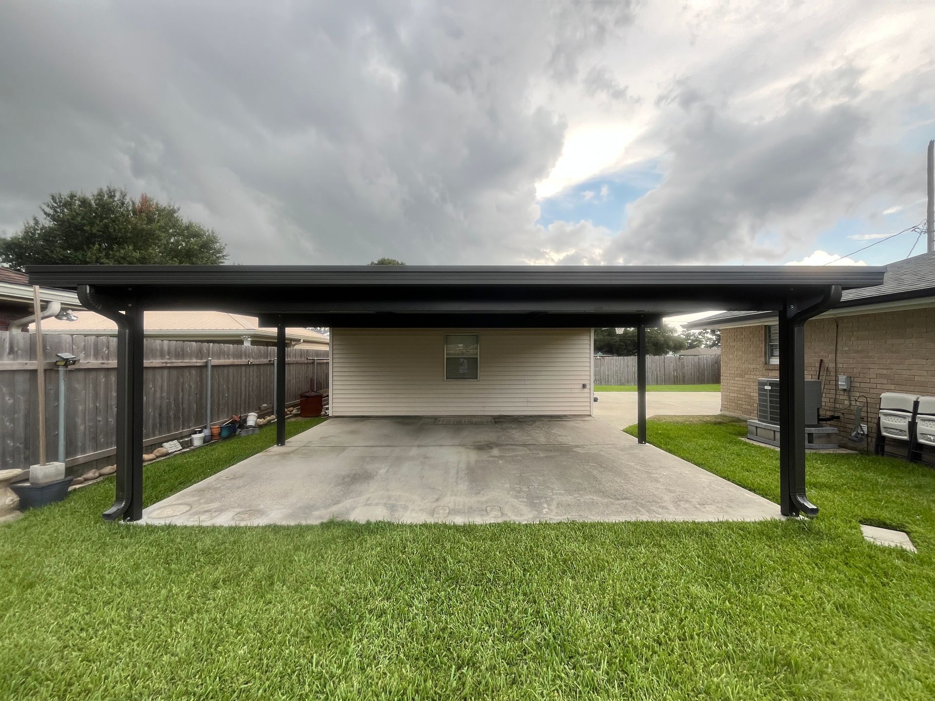 A modern black metal carport stands over a concrete driveway next to a residential house and lawn under a cloudy sky.