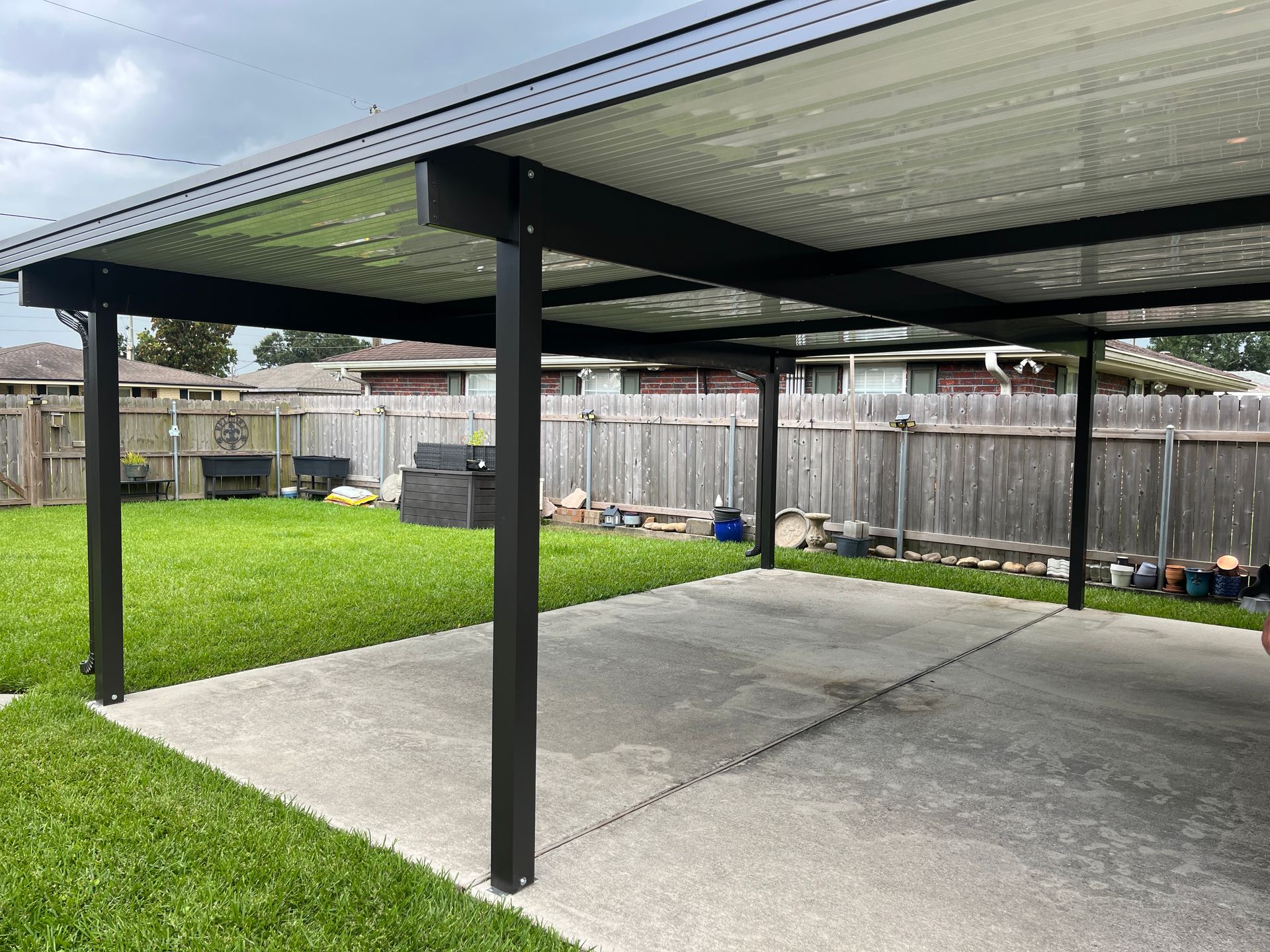 A black-framed patio cover over a concrete slab in a grassy backyard with a wooden fence in the background.