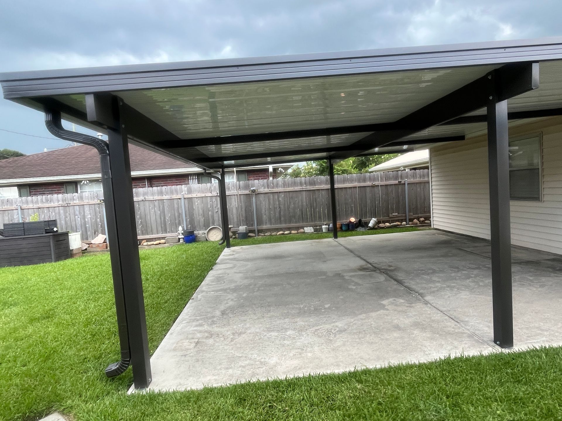 A covered patio with a dark metal frame and roof, attached to a house over a concrete driveway next to a grassy lawn.